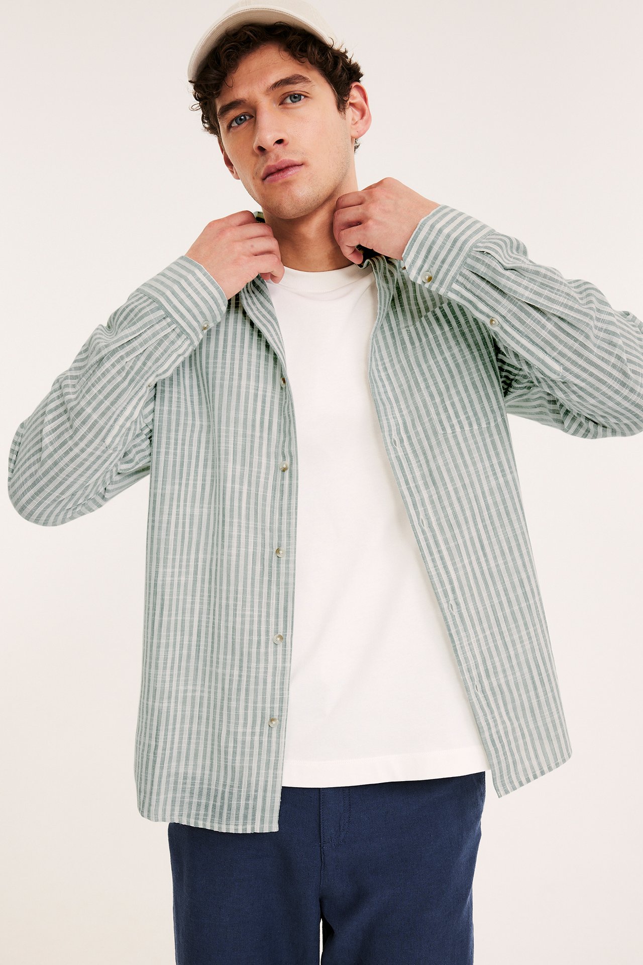 Mies päällä avoin vaaleanvihreä raidallinen puuvillapaita, valkoinen t-paita, siniset housut ja beige lippalakki. Edestä.