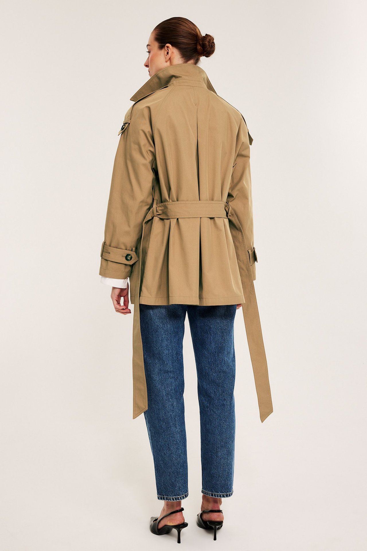 Nainen takaa, yllään beige leveä trenssitakki vyöllä, siniset farkut ja mustat korkokengät.