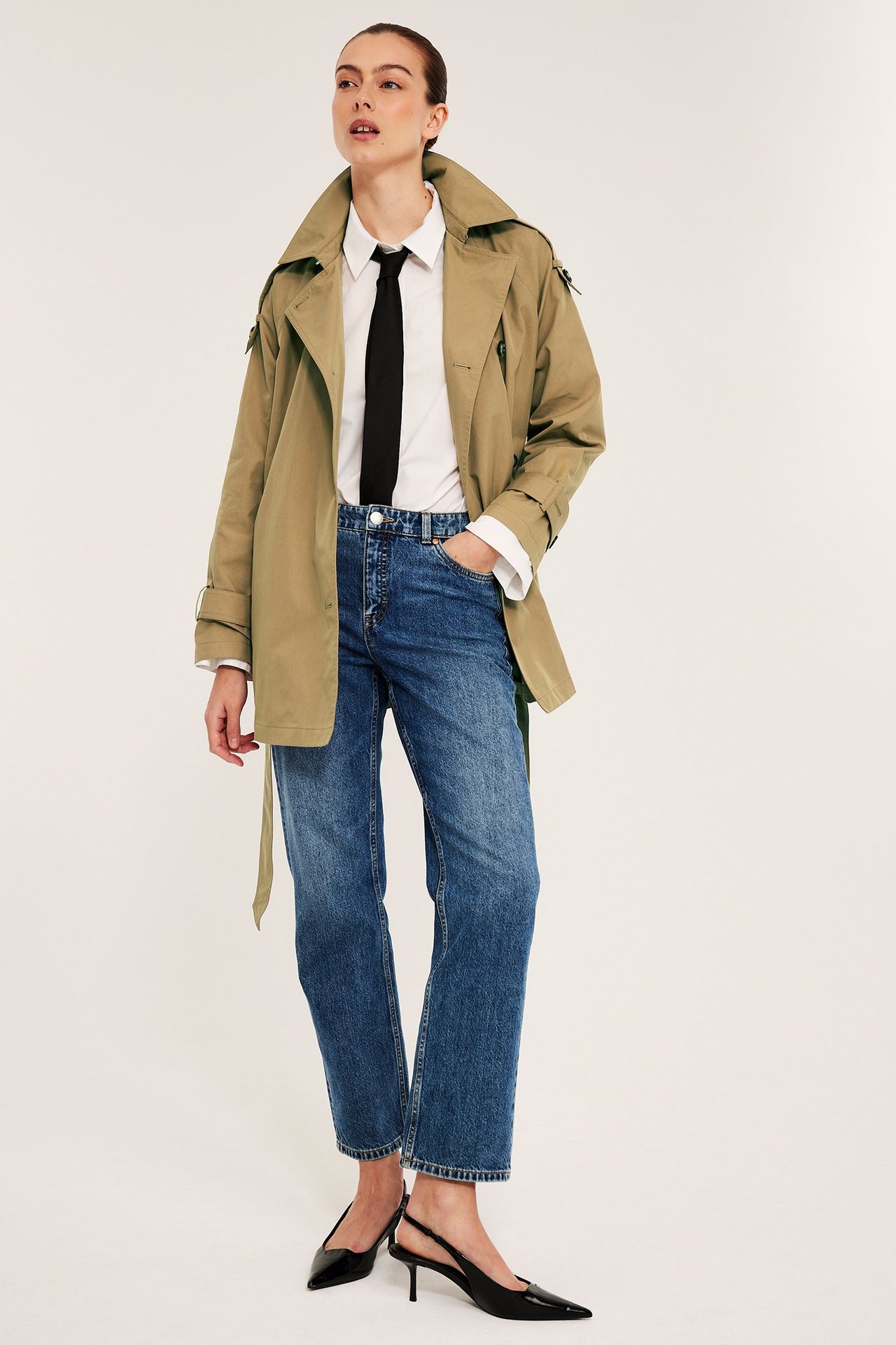 Edestä: Nainen beigessä leveässä trenssitakissa, valkoinen paita, musta solmio, siniset farkut ja mustat avokantakengät.