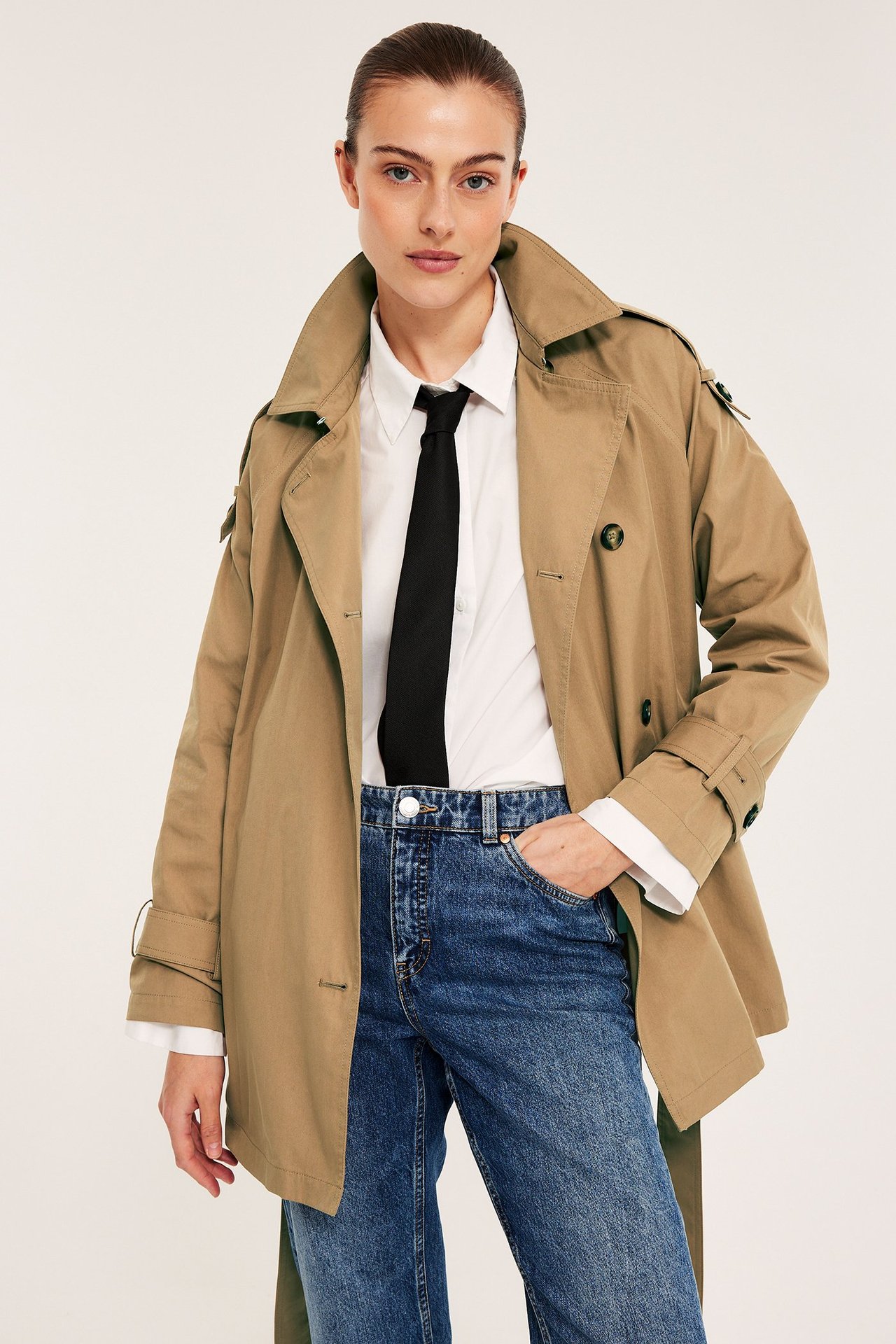 Edestä: Nainen beigessä trenssitakissa, valkoisessa paidassa, mustassa solmiossa, sinisissä farkuissa.