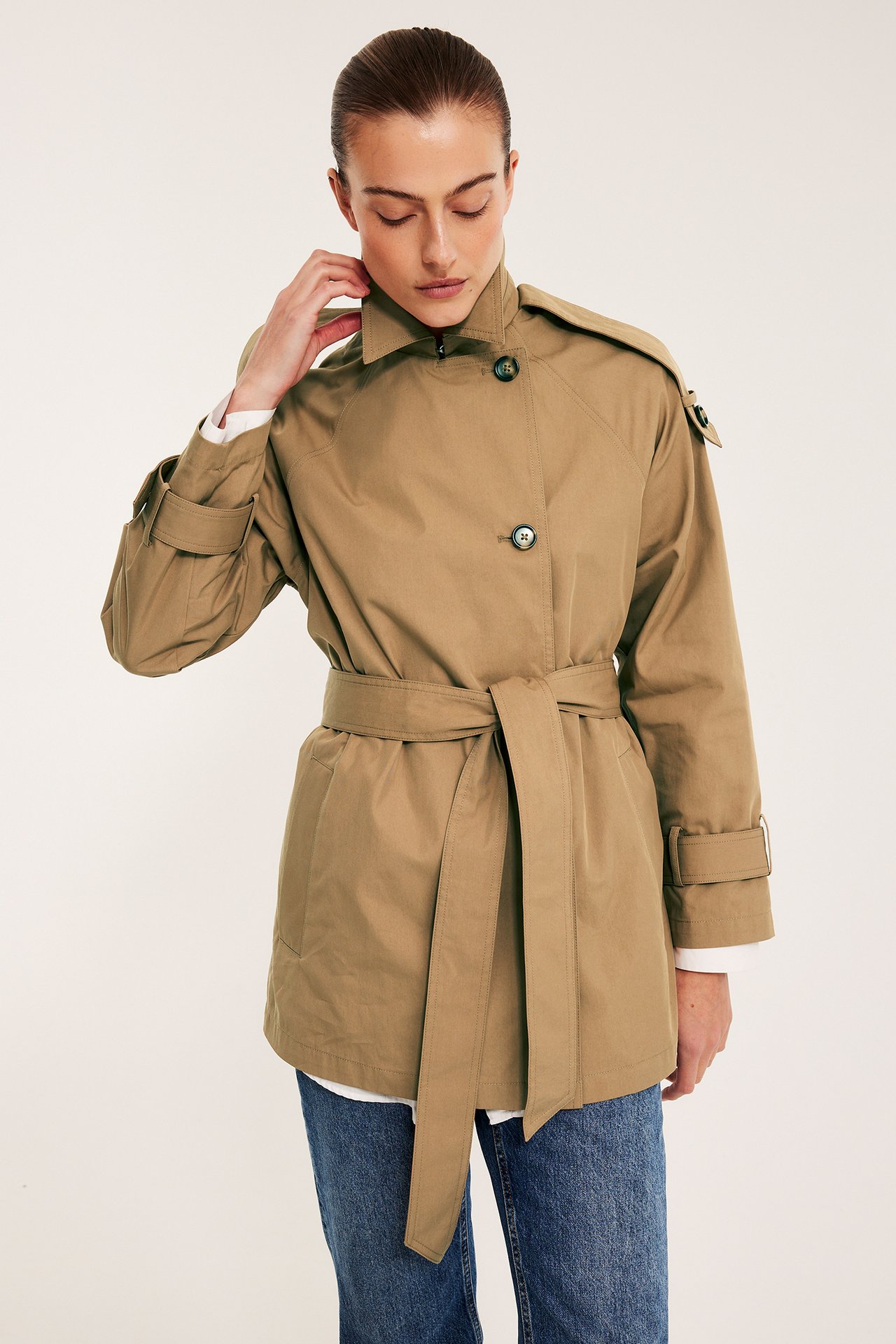 Nainen edestäpäin beigessä lyhyessä trenssitakissa vyöllä, valkoisen paidan ja sinisten farkkujen päällä.