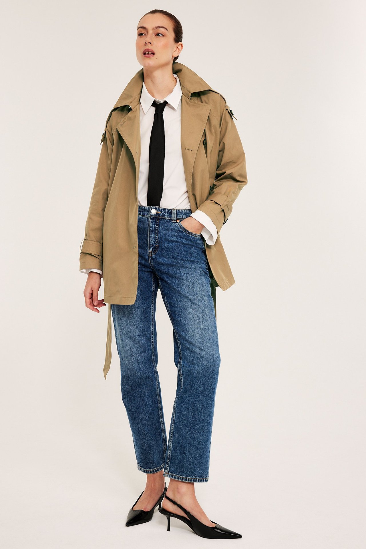 Kvinne forfra i beige trenchcoat over hvit skjorte, svart slips, blå jeans og svarte hæler.