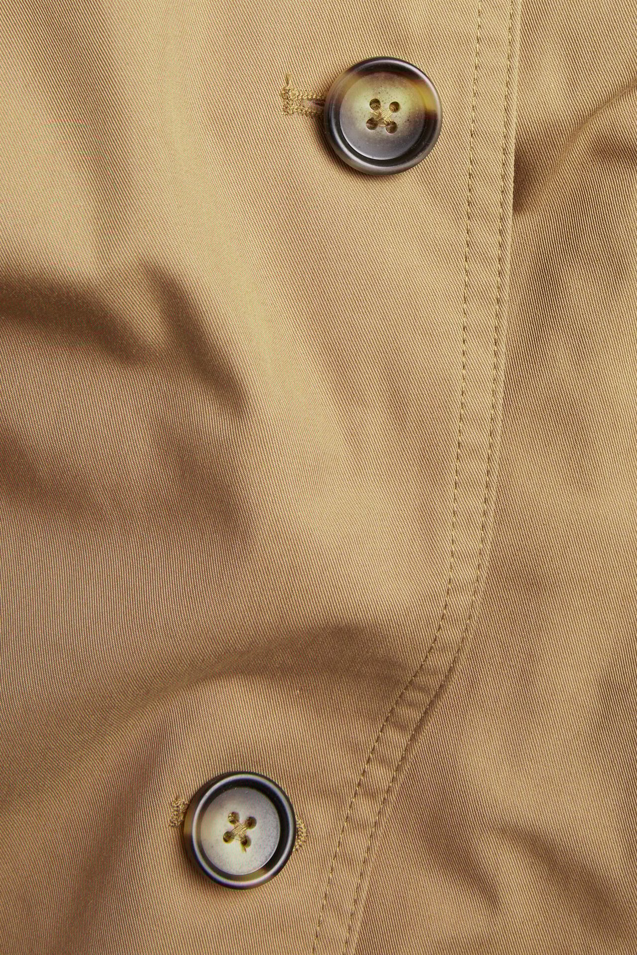 Zbliżenie beżowego trenczu z dwoma brązowymi, wzorzystymi guzikami i widocznym szwem.