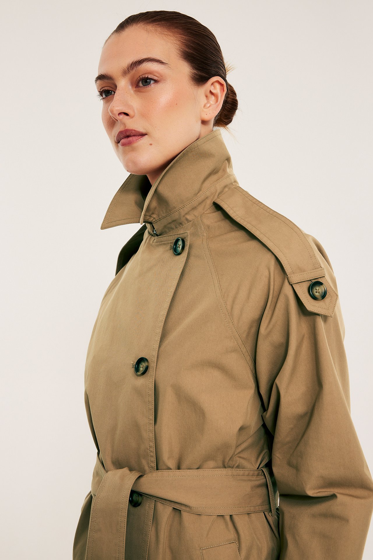 Nærbilde, sidevisning av en kvinne i en vid beige trenchcoat med knytebelte og mørke knapper.