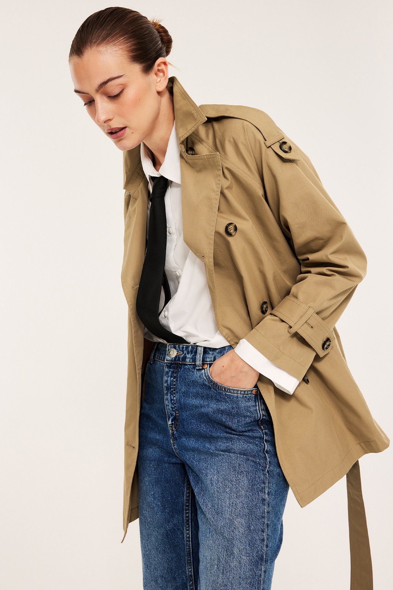 Forfra: kvinne i åpen beige mellomlang trenchcoat, hvit skjorte, svart slips og blå jeans.
