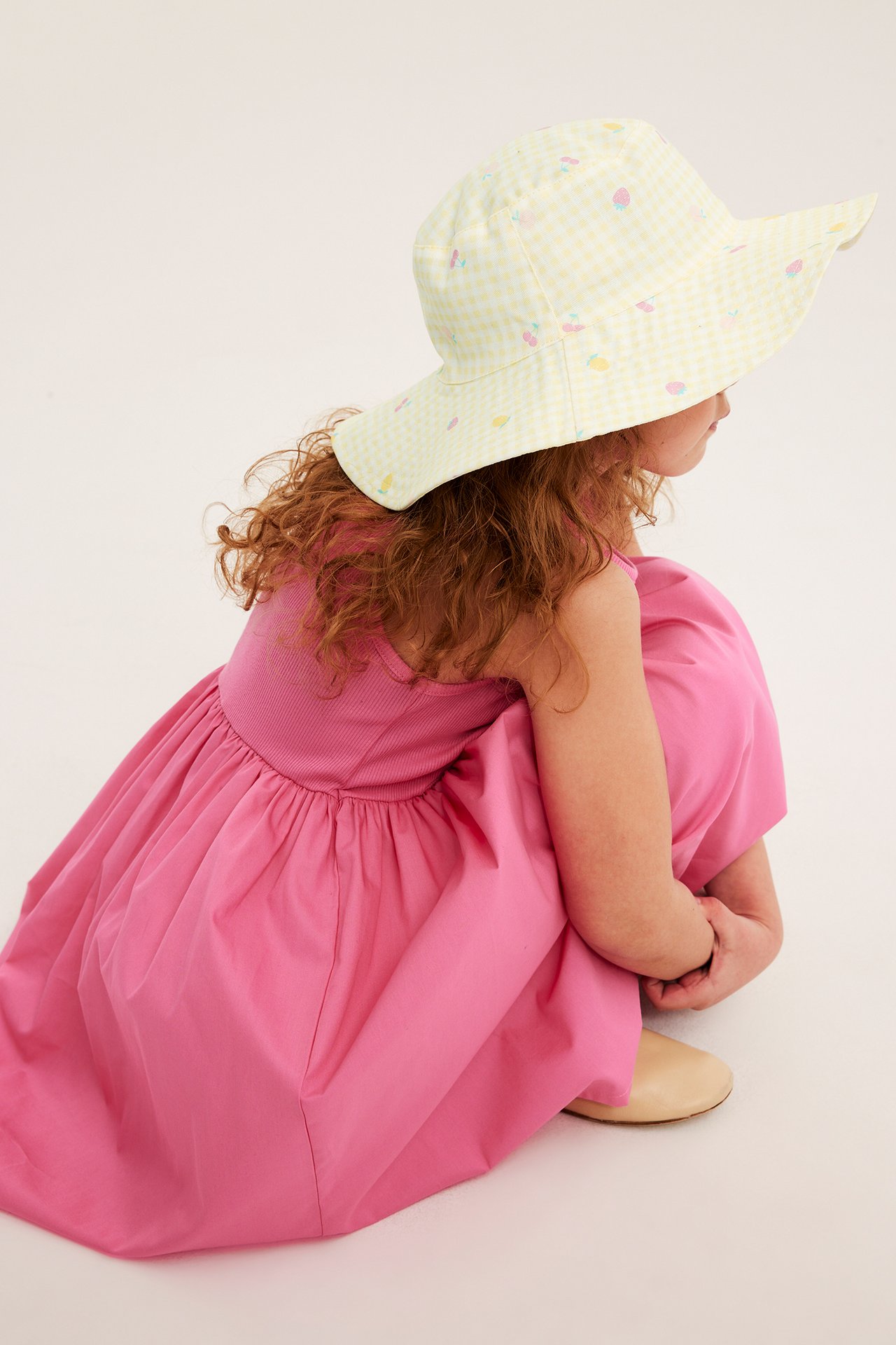 Takaa: Lapsi yllään vaaleankeltainen ruudullinen aurinkohattu hedelmäkuviolla ja pinkki mekko.