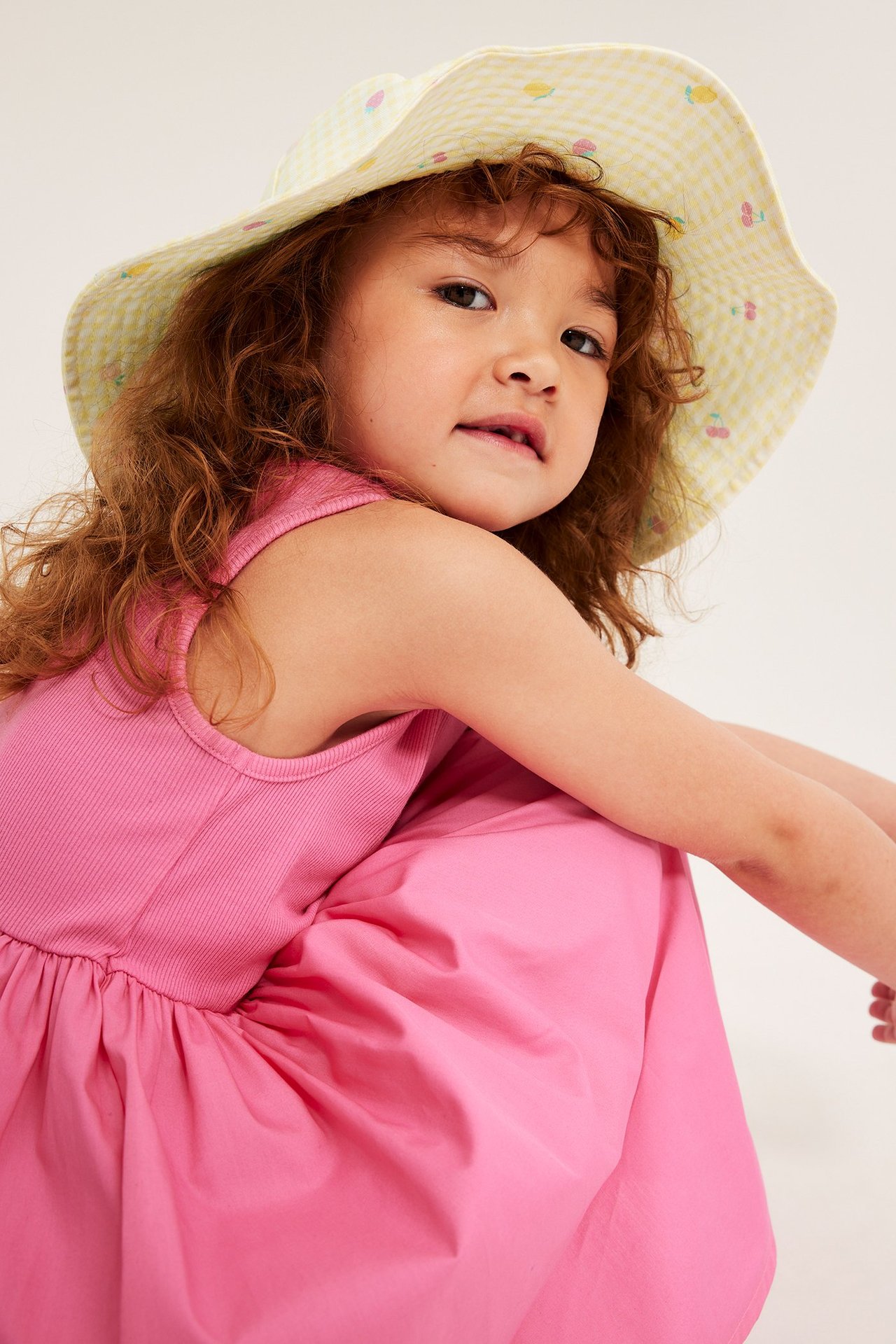 Lapsi vaaleankeltaisessa ruudullisessa aurinkohatussa hedelmäprintillä ja vaaleanpunaisessa mekossa, edestä.