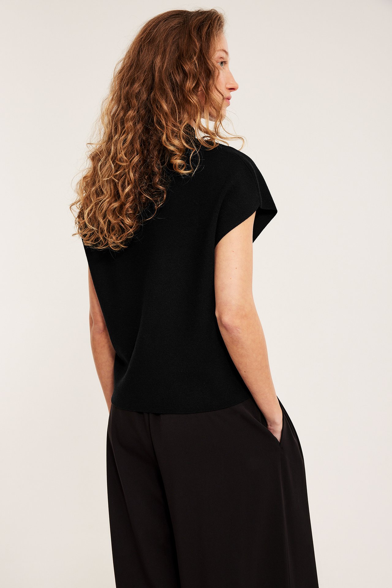 Side-/bakfra av kvinne i svart strikket vest med krage, korte ermer og svarte bukser.