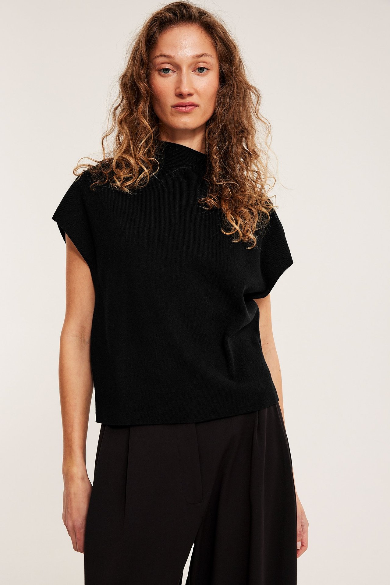 Edestä näkymä: nainen käyttää mustaa neulottua liiviä kauluksella ja mustia leveälahkeisia housuja.