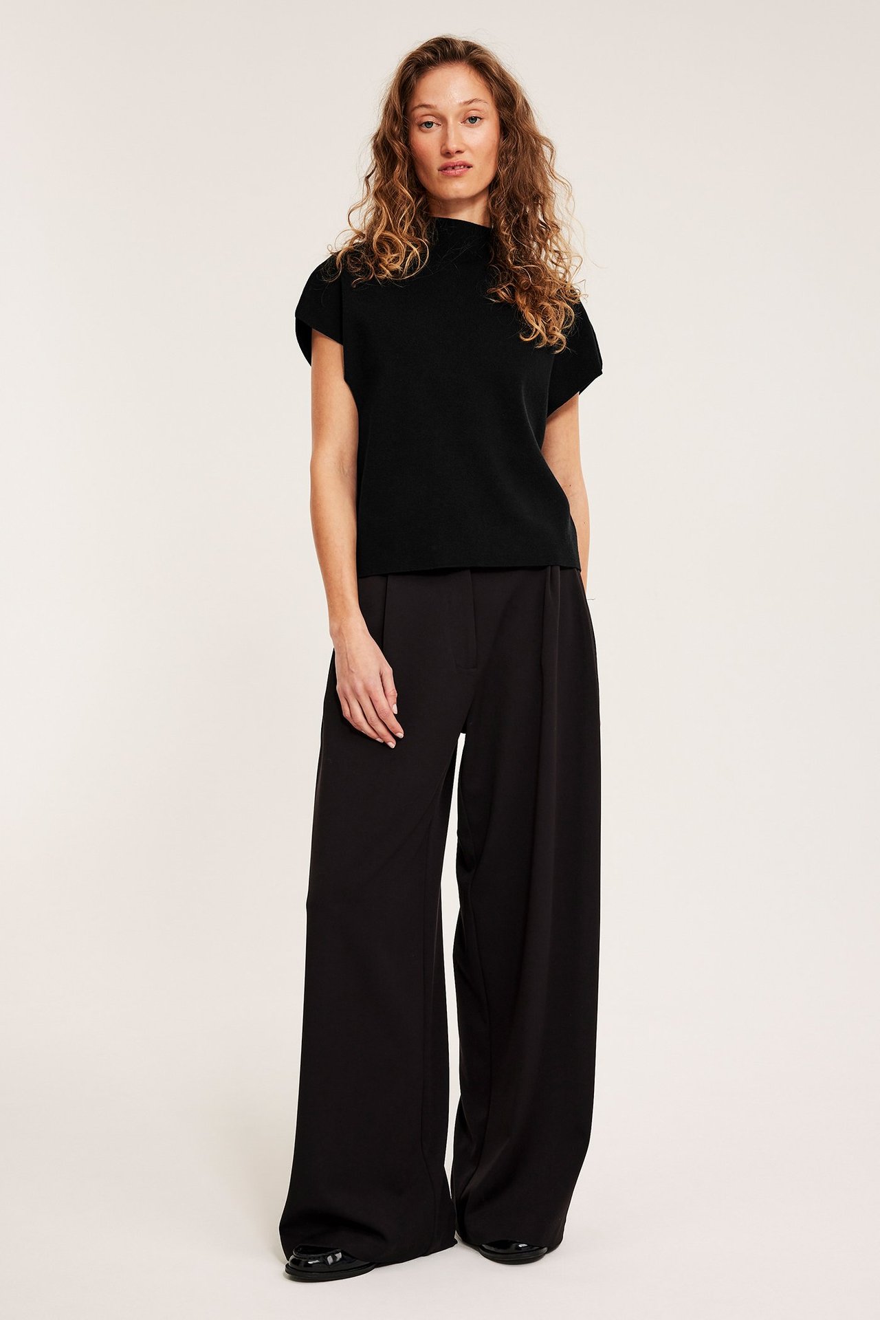 Front, kvinne i svart strikket høyhalset vest, sorte vide bukser og sorte flate sko.