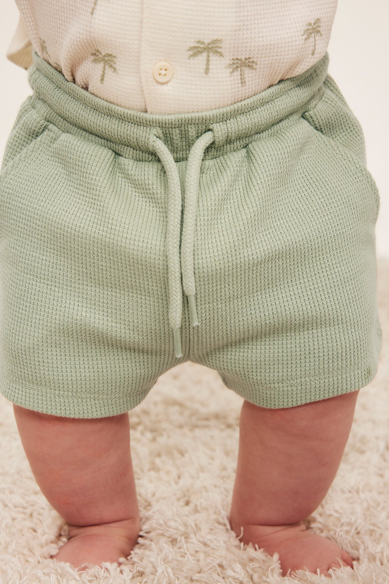 Närbild framifrån på baby i ljusgröna våfflade shorts med dragsko och vit tröja med palmträdsmönster.