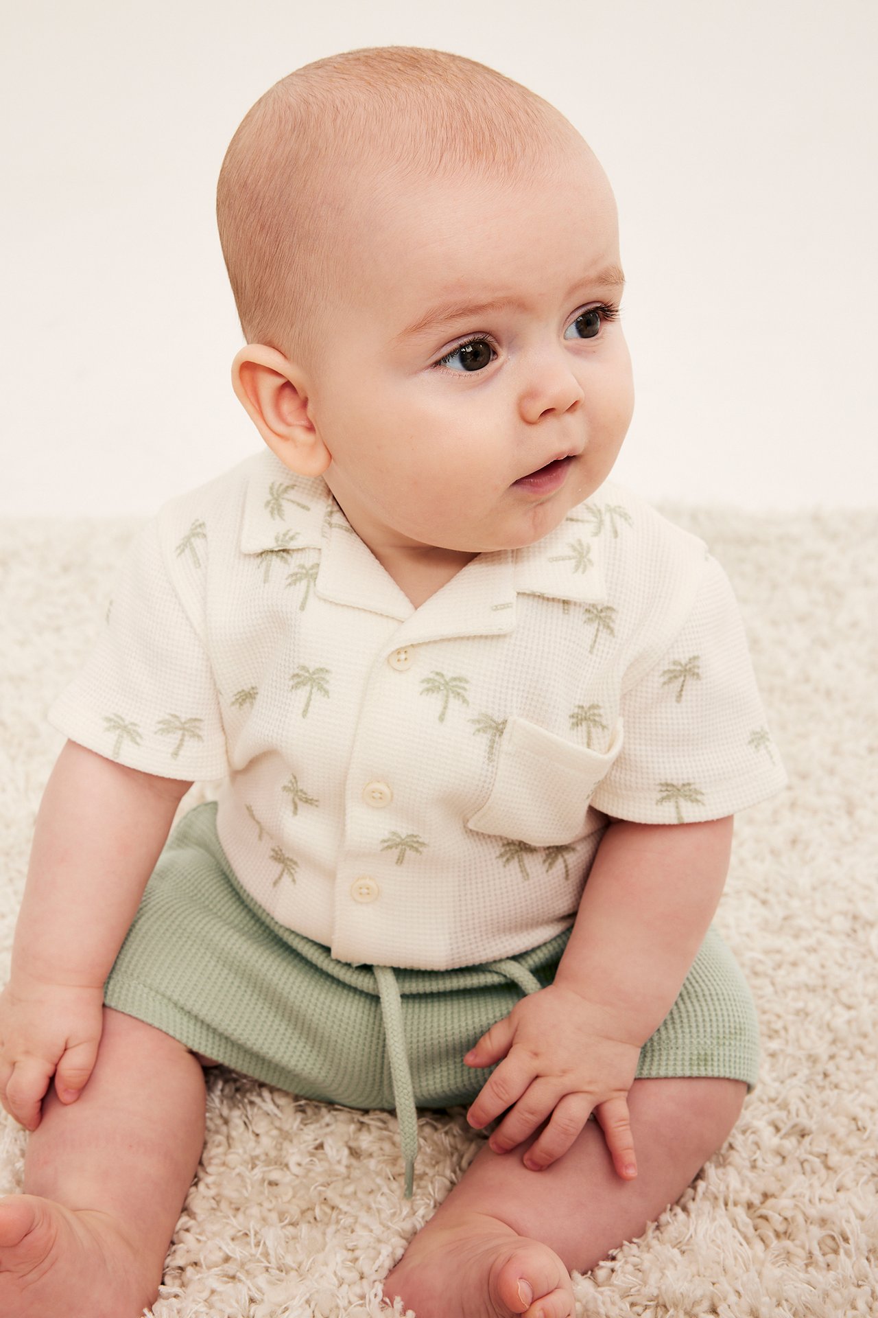 Framifrån, baby i ljusgröna våfflade shorts med dragsko och en skjorta med palmträd.
