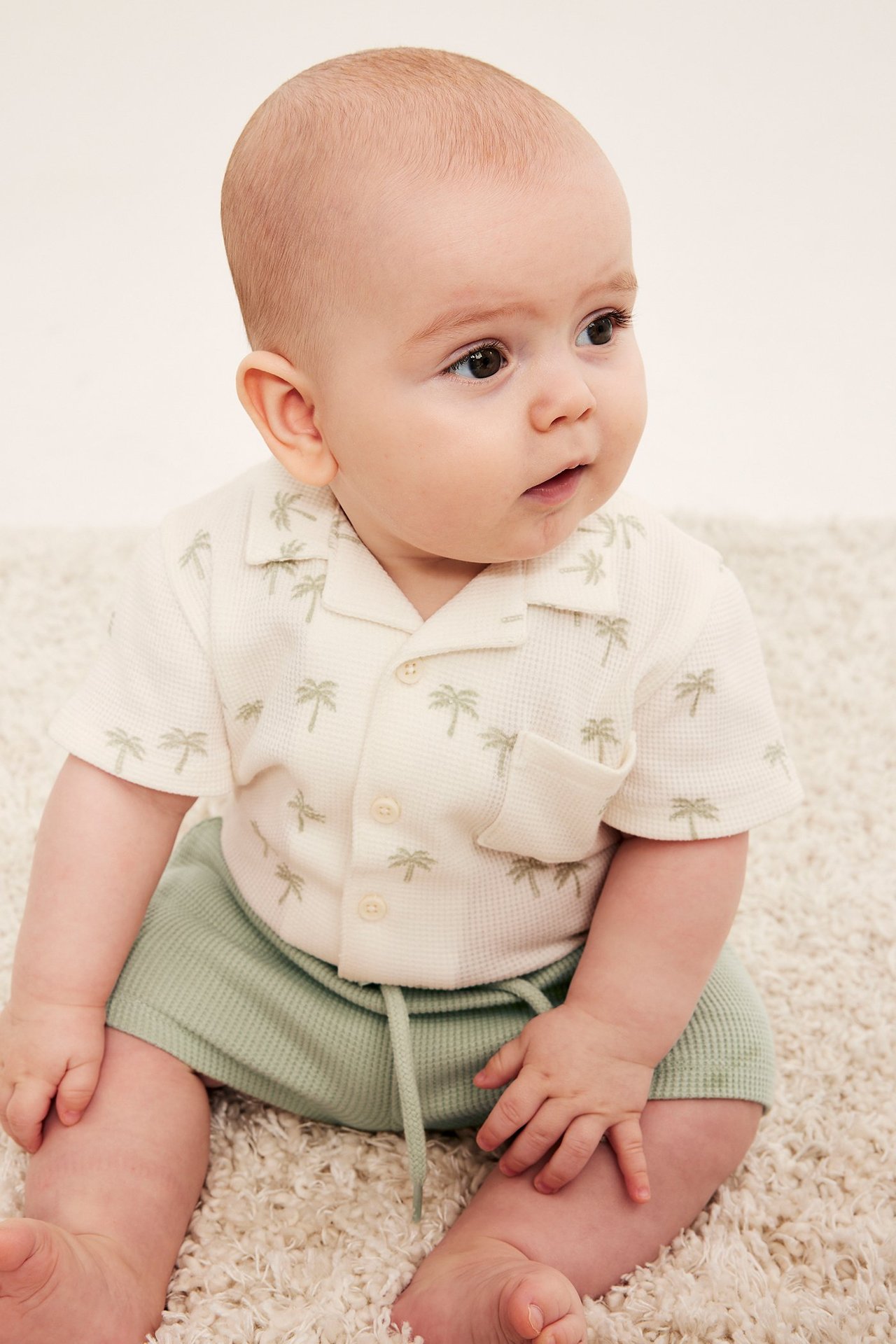 Framifrån, baby i ljusgröna våfflade shorts med dragsko och en skjorta med palmträd.