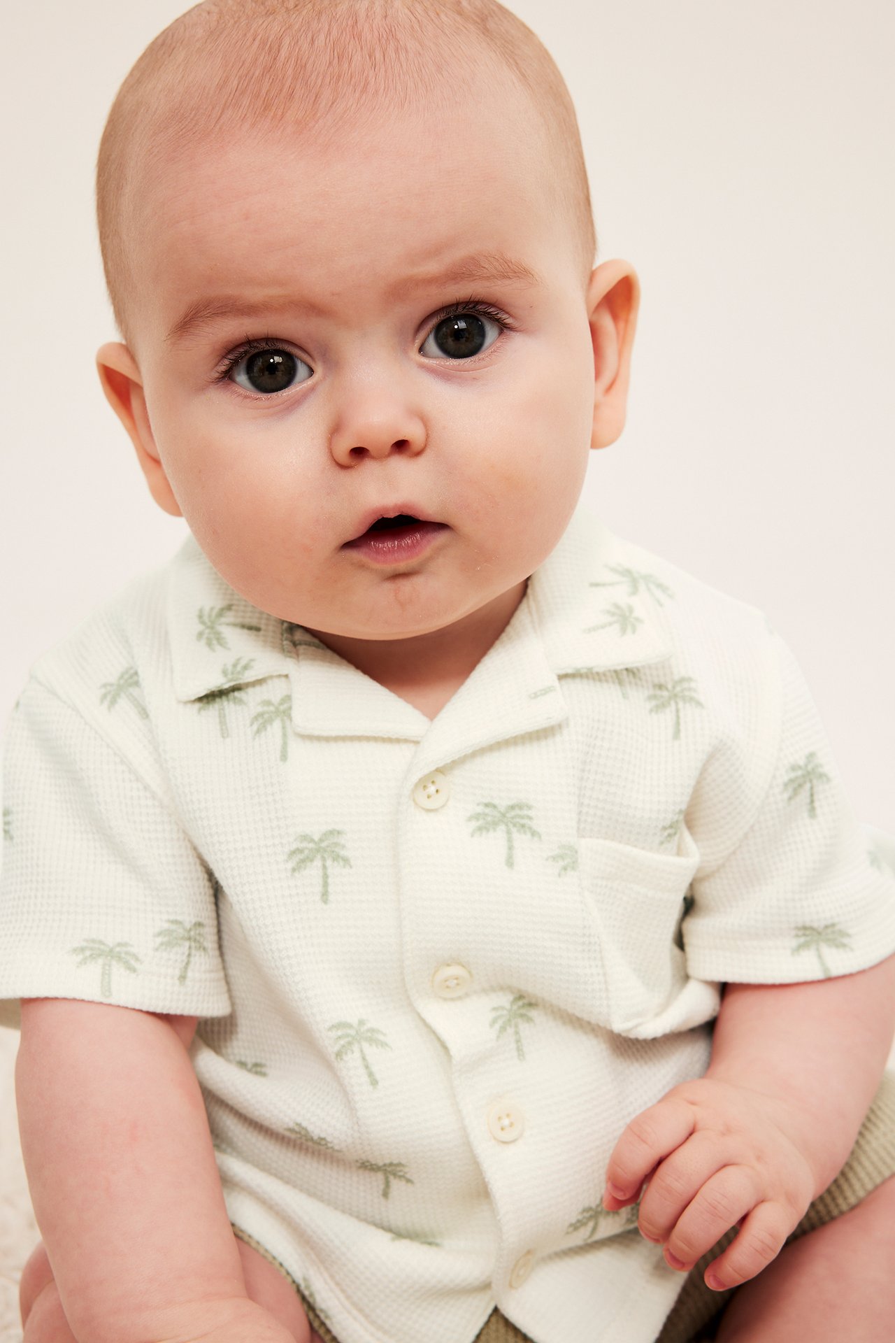 Närbild på baby i offwhite våfflad kortärmad skjorta med palmträdsmönster och bröstficka.