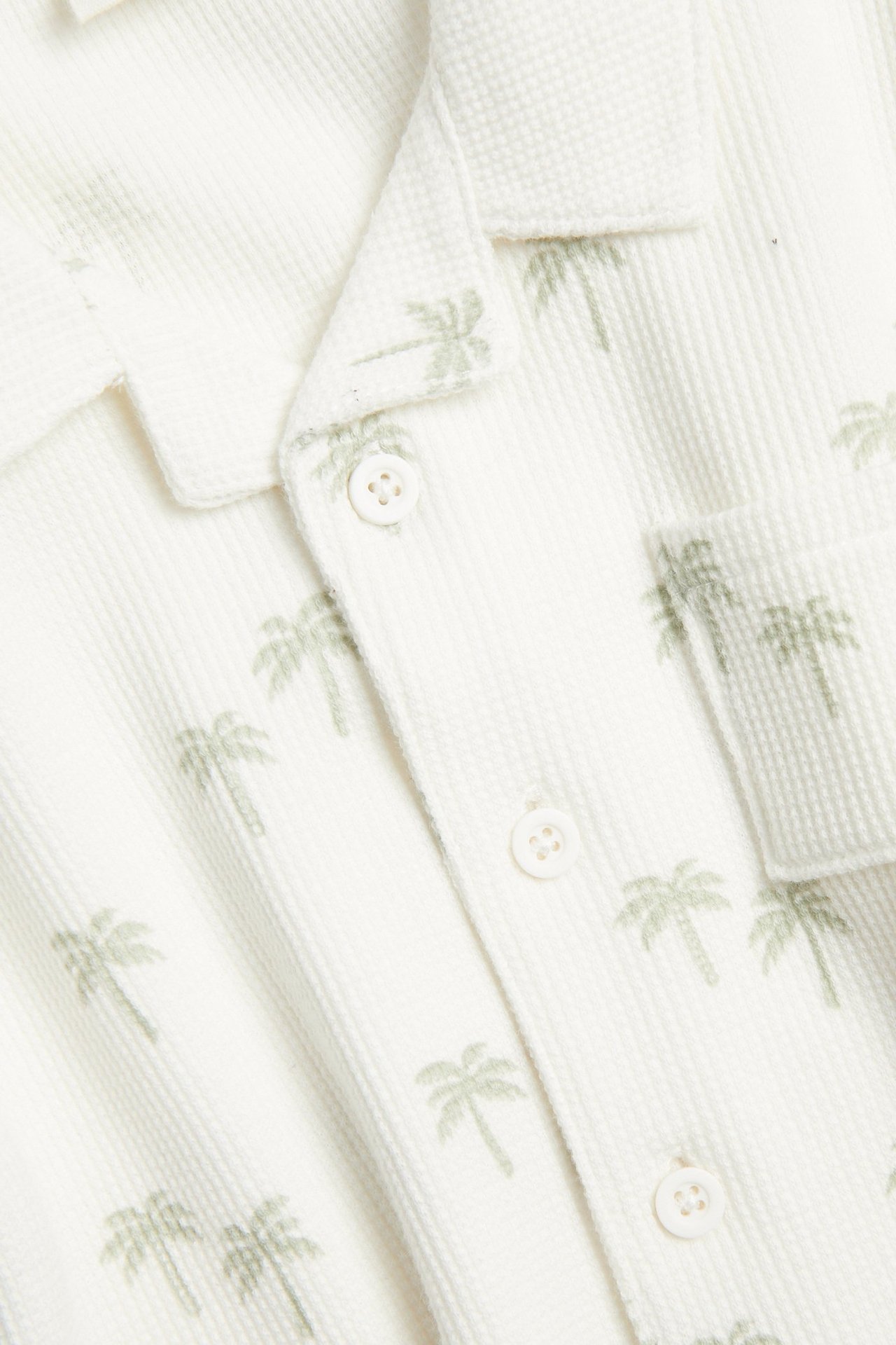 Nærbilde forfra av en babys offwhite kortermet vaffelskjorte med lysegrønne palmer og knapper.