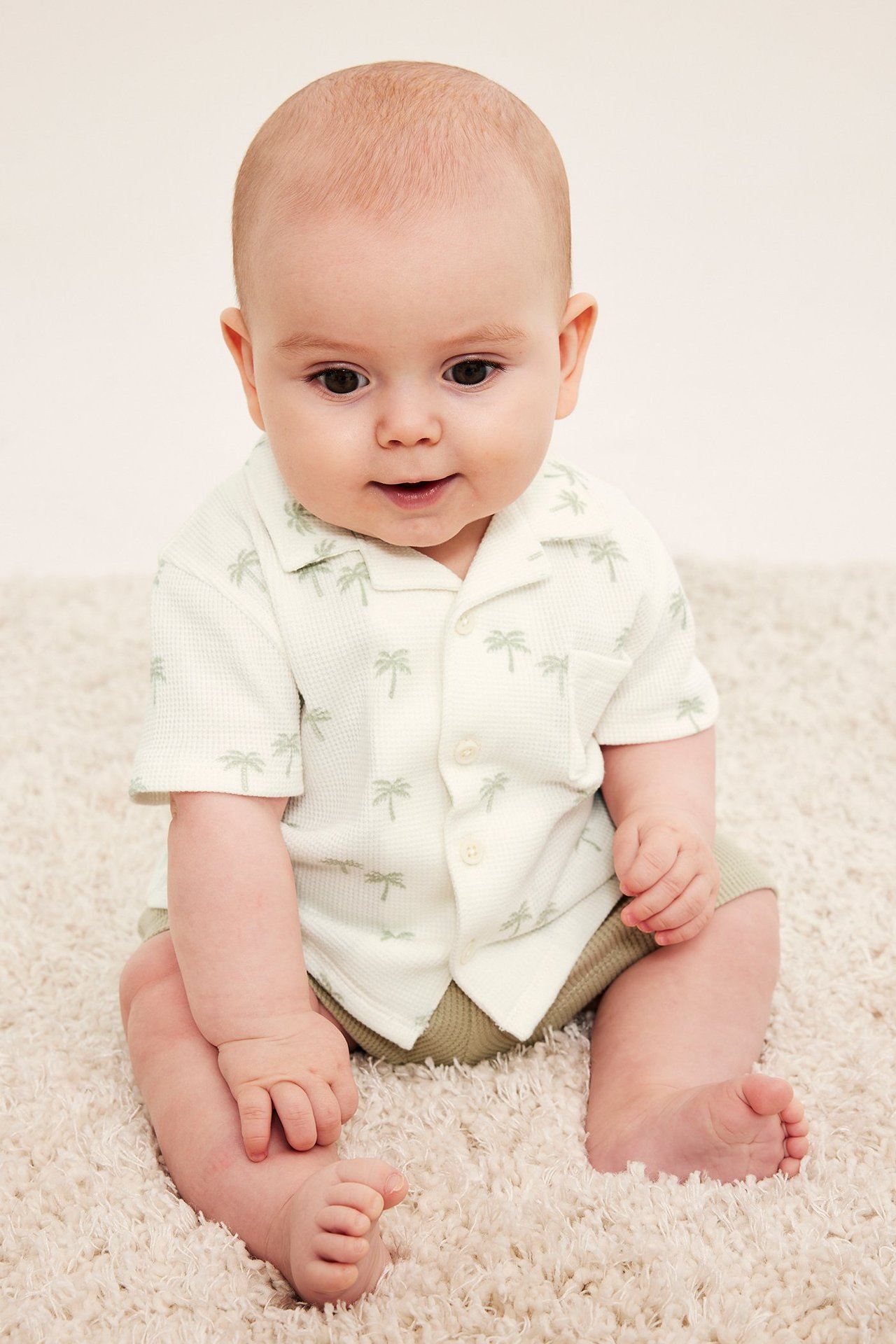 Framifrån: Baby bär en offwhite våfflad skjorta med palmtryck, ficka och khaki shorts.