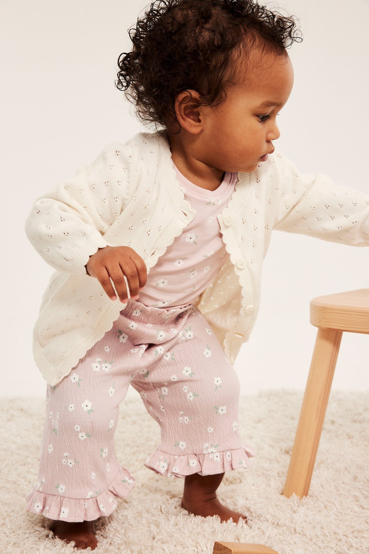 Framsida på baby i rosa småblommiga byxor med volangkanter, en rosa topp och en vit stickad kofta.