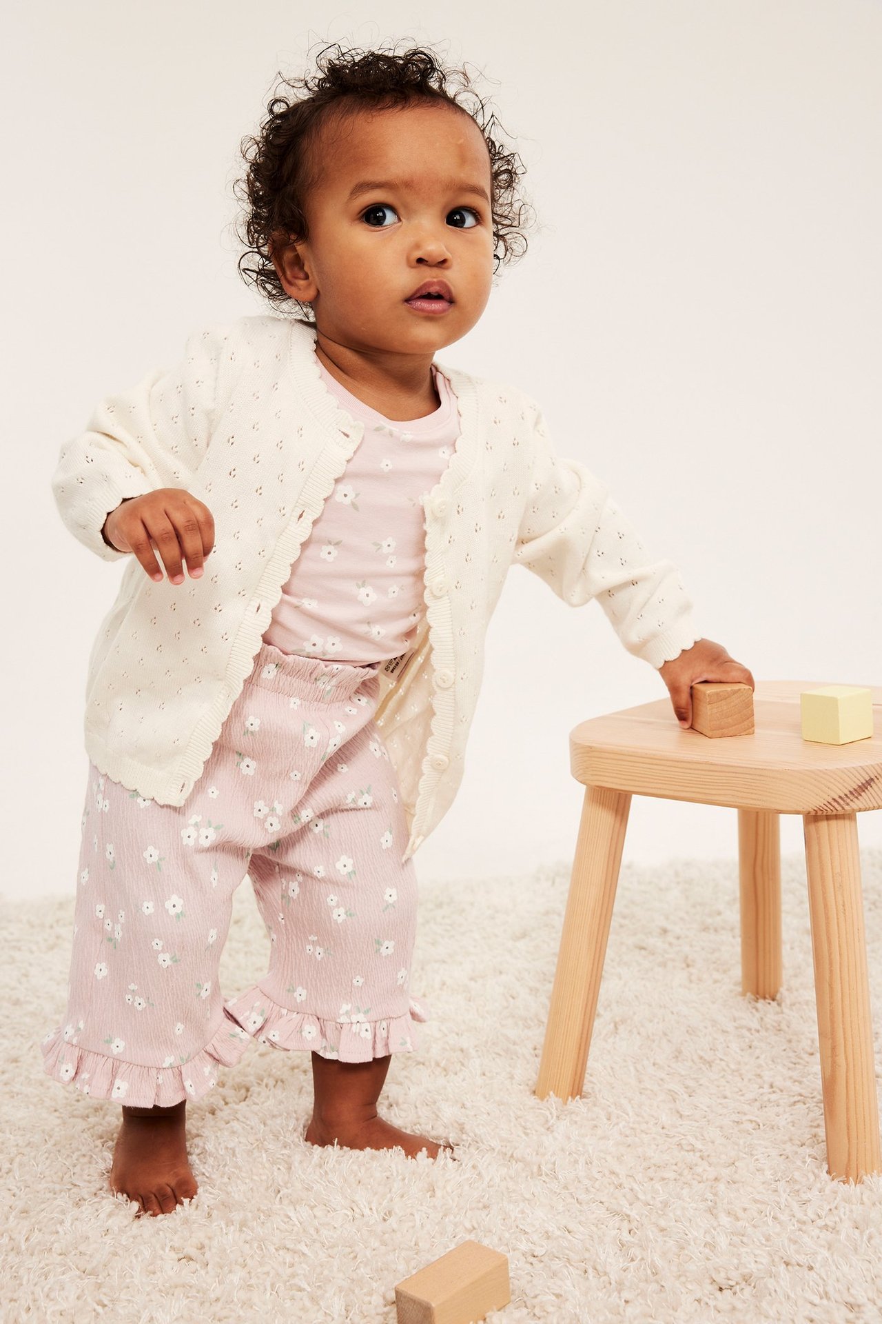 Edestäpäin, vauva vaaleanpunaisissa kukkakuvioisissa puuvillahousuissa, joissa röyhelöt, pinkki t-paita ja kermanvärinen neuletakki.