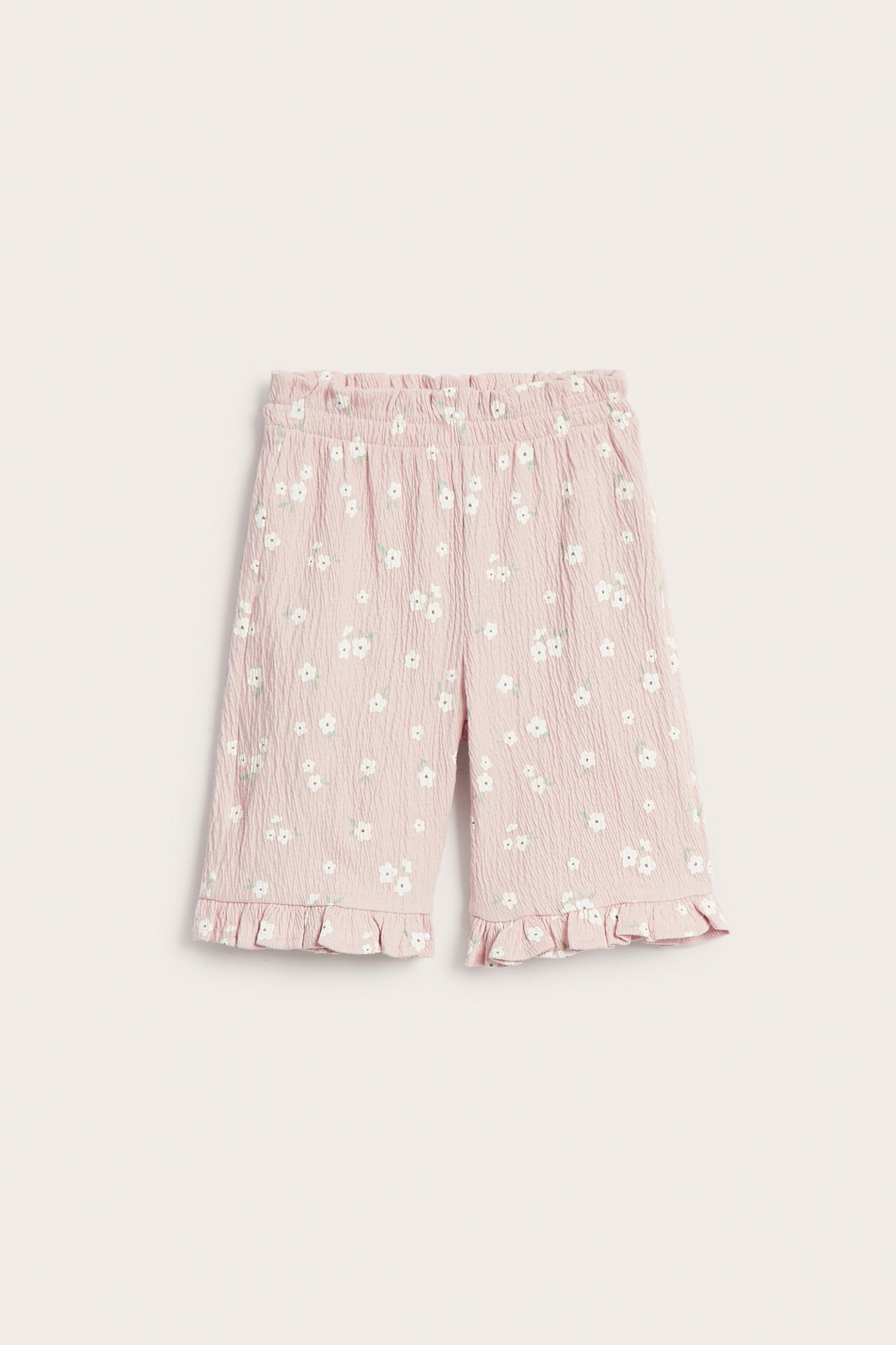 Frontvisning av babyens rosa bomullskreppbukse med små blomster, elastisk volangmidje og volanger ved bena.