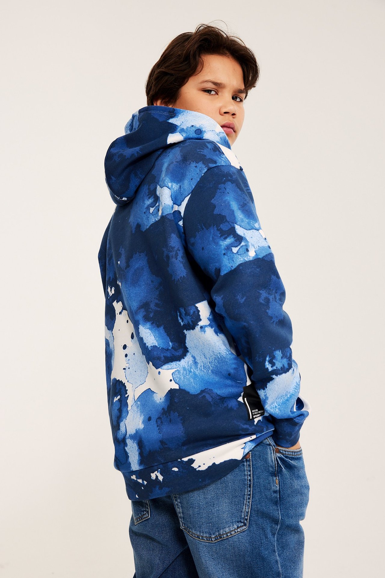 Widok z boku i z tyłu: Dziecko w niebiesko-białej wzorzystej bluzie z kapturem i niebieskich jeansach.