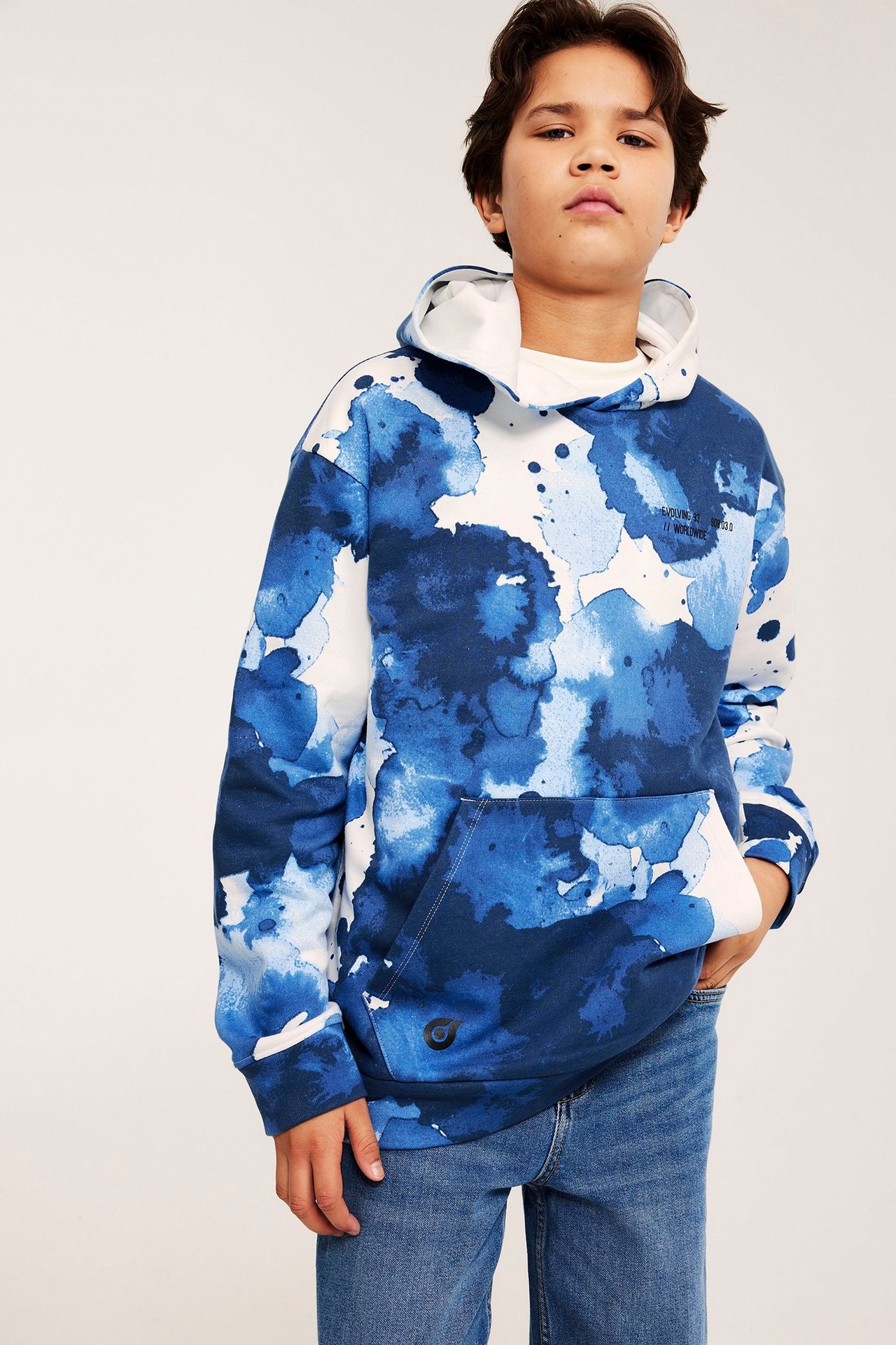 Widok z przodu: Chłopiec w białej bluzie z kapturem, niebieskim akwarelowym wzorem, kieszenią i dżinsach.