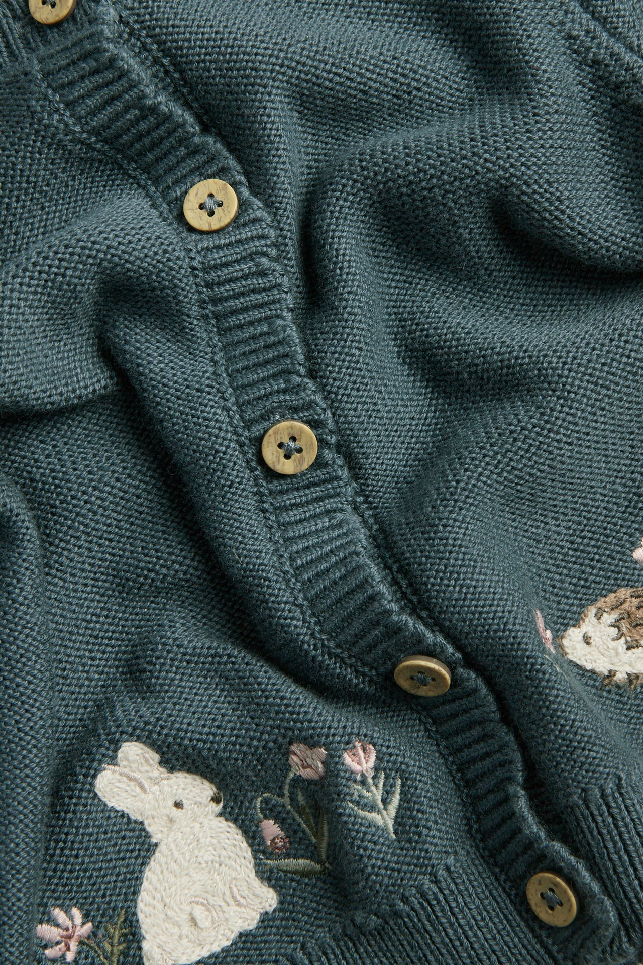Nærbilde av en mørk turkis strikket cardigan med treknapper og brodert kanin, pinnsvin og blomster.
