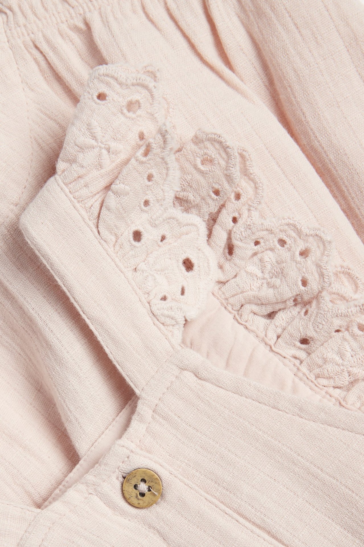 Nærbilde forfra av lyserosa vevde bomullsselebukser med blondevolanger på stroppene og en treknapp.