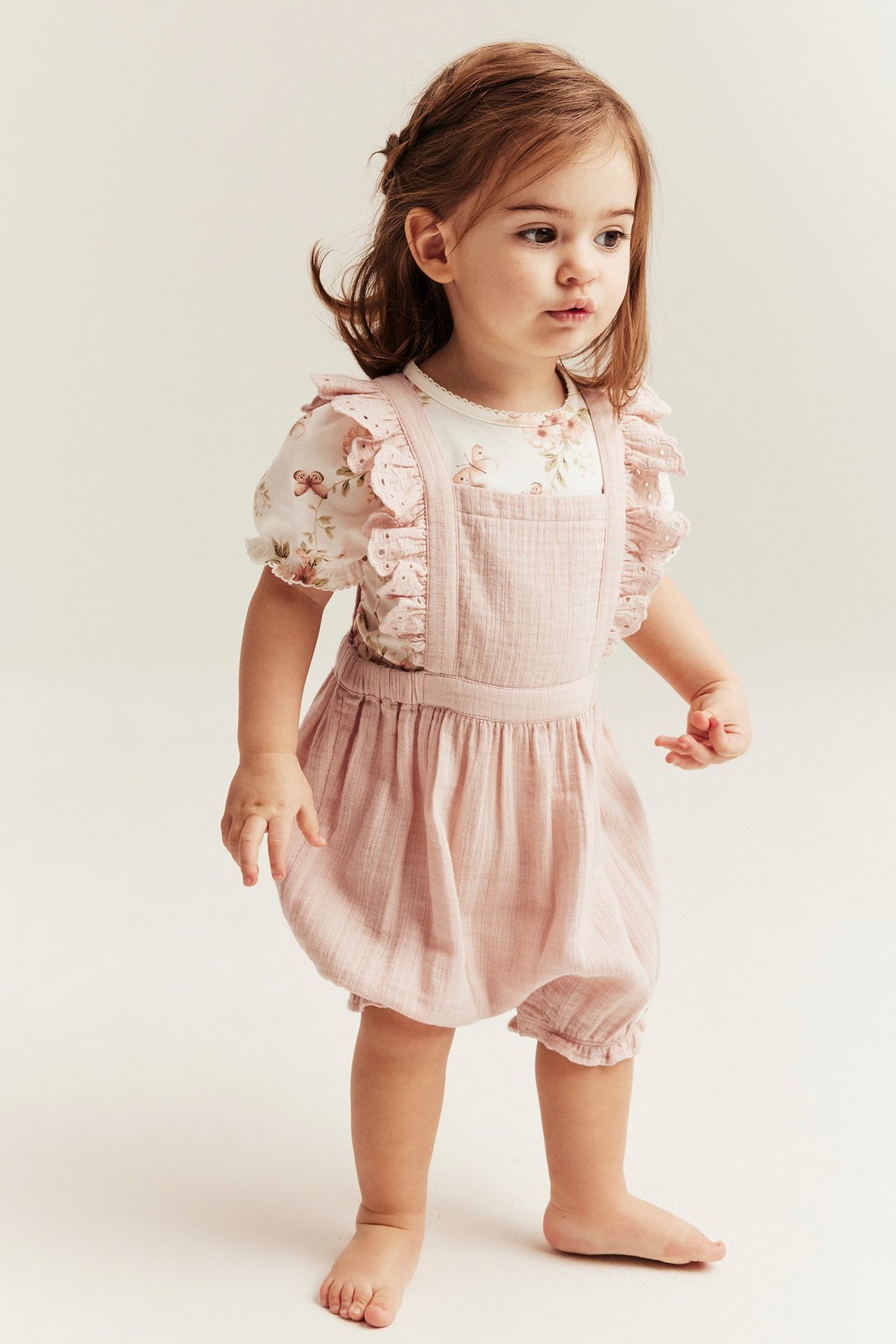 Widok z przodu dziewczynki w jasnoróżowych szelkach z falbankami na białym topie w kwiaty.