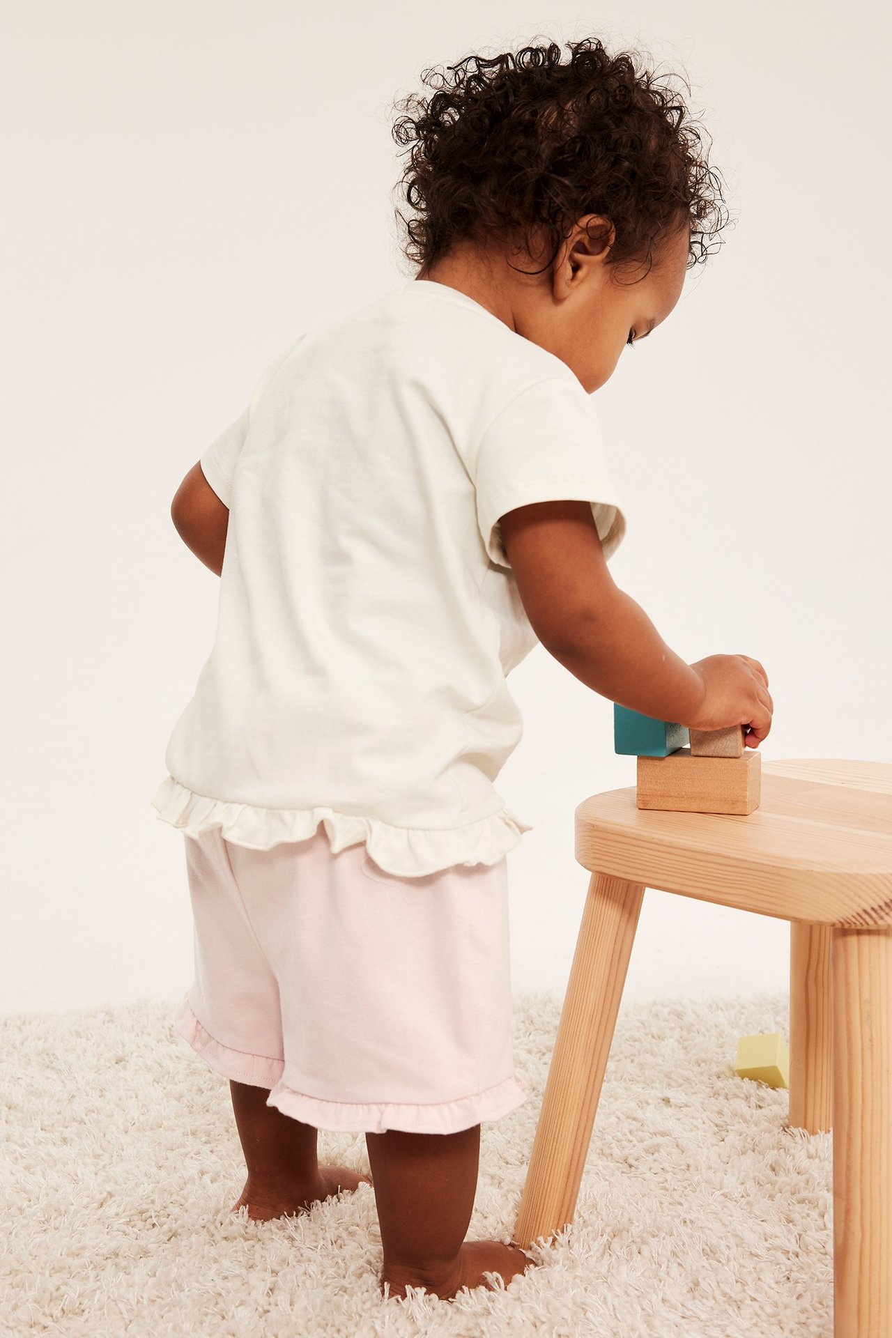 Niemowlę, tyłem, w różowych, jerseyowych szortach z falbankami i ecru T-shircie, bawi się klockami.