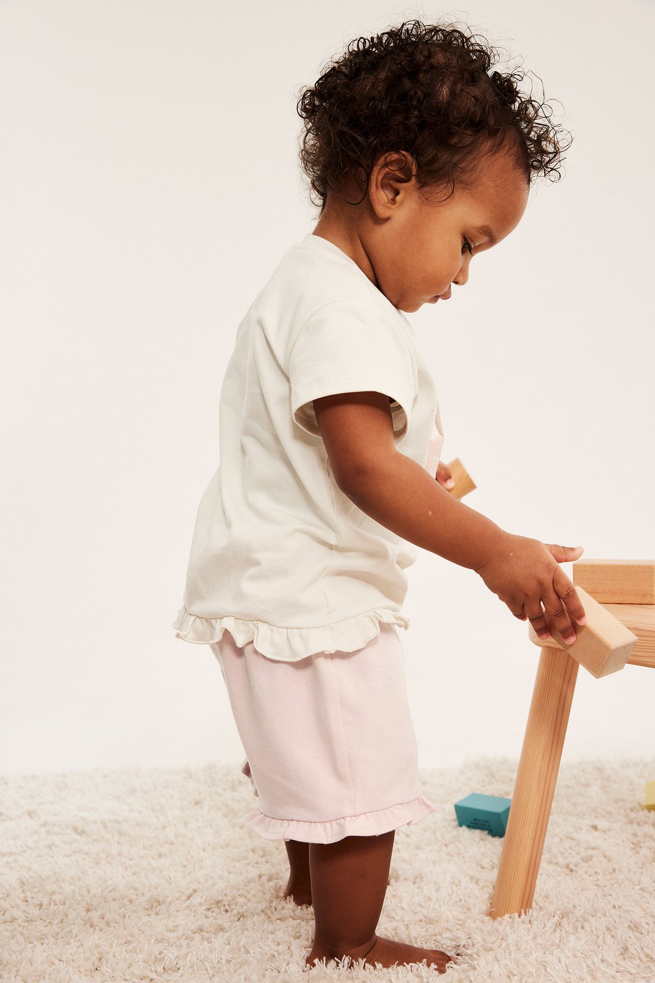 Vauva sivulta, vaaleanpunaiset röyhelöshortsit ja kermanvärinen t-paita, leikkii puupalikoilla.