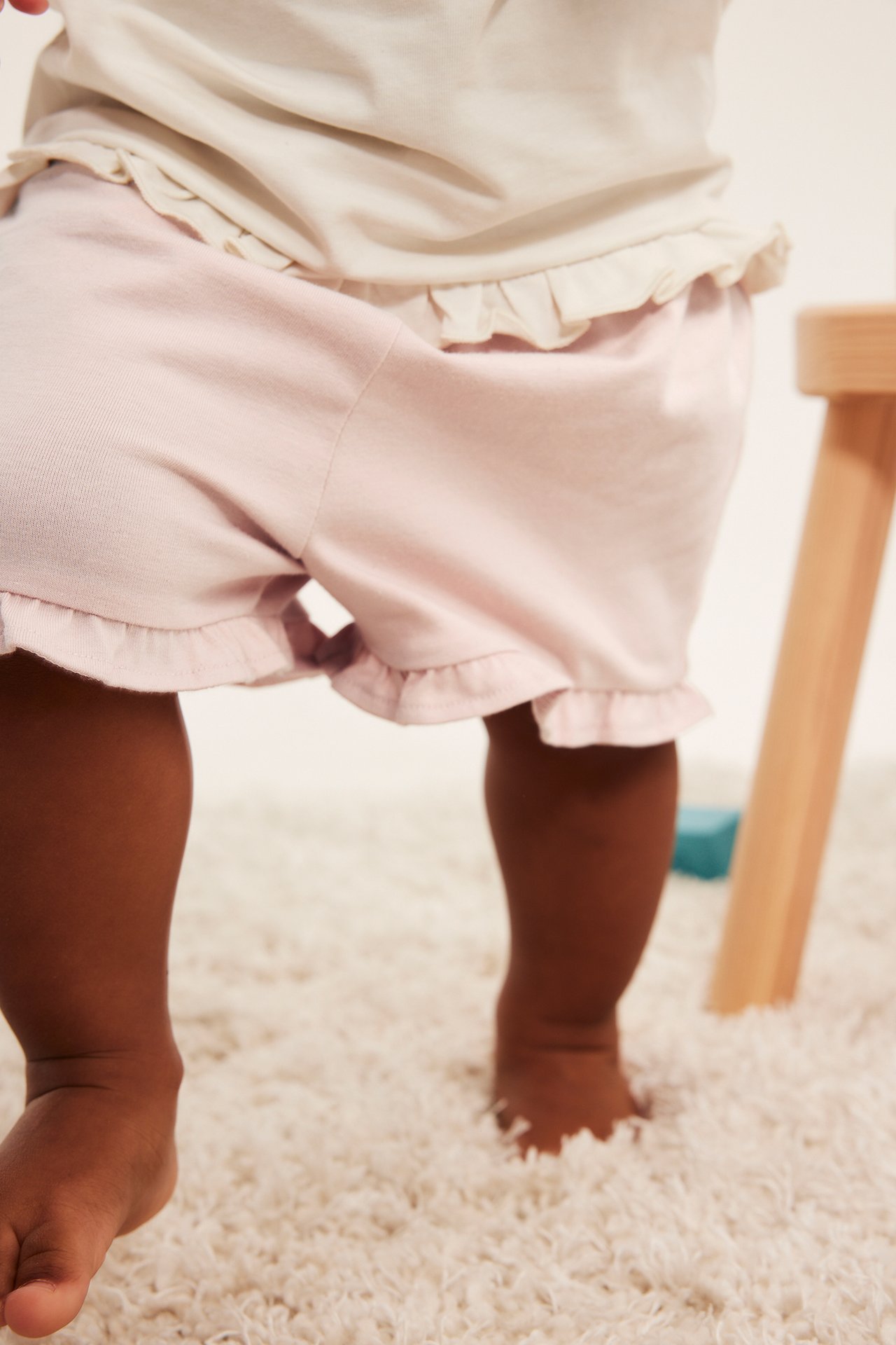 Edestä: vauva yllään vaaleanpunaiset joustavat shortsit röyhelöreunalla ja vaalea toppi.