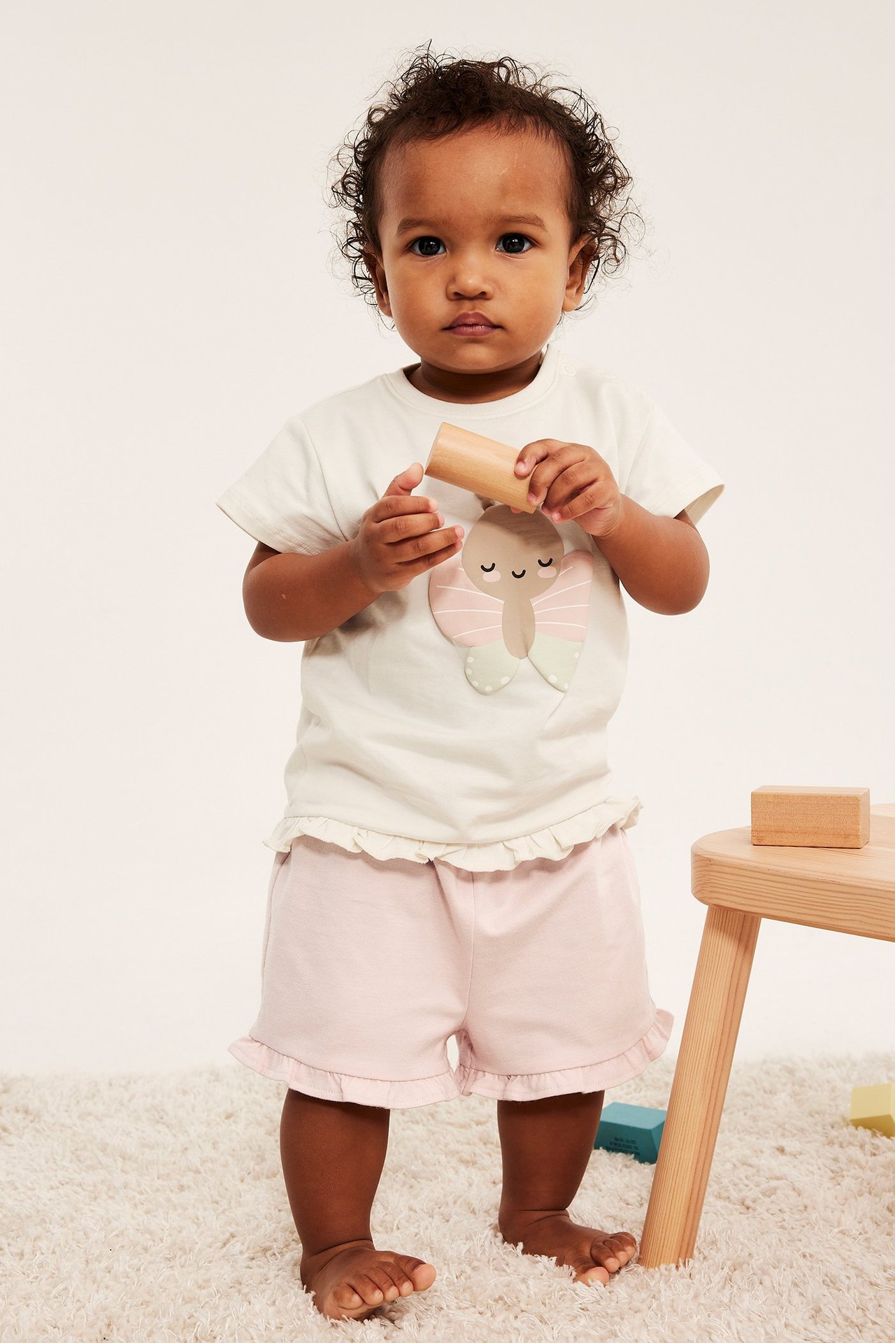Różowe szorty dżersejowe z falbankami na dziecku, z jasnobeżową koszulką w motyle, trzyma drewniany klocek.
