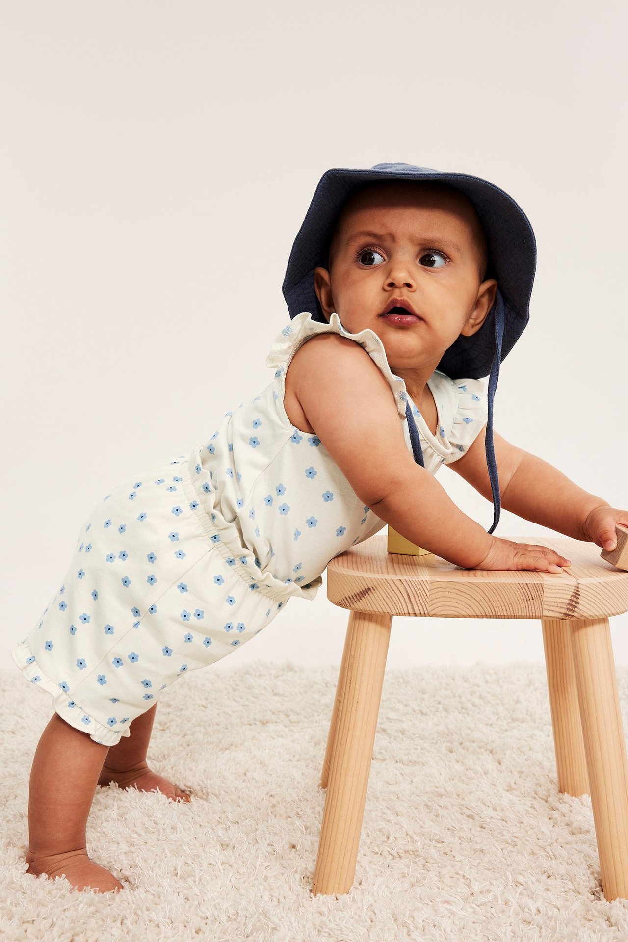 Widok z przodu: Niemowlę w kremowych, kwiecistych szortach z falbankami i granatowym kapeluszu.