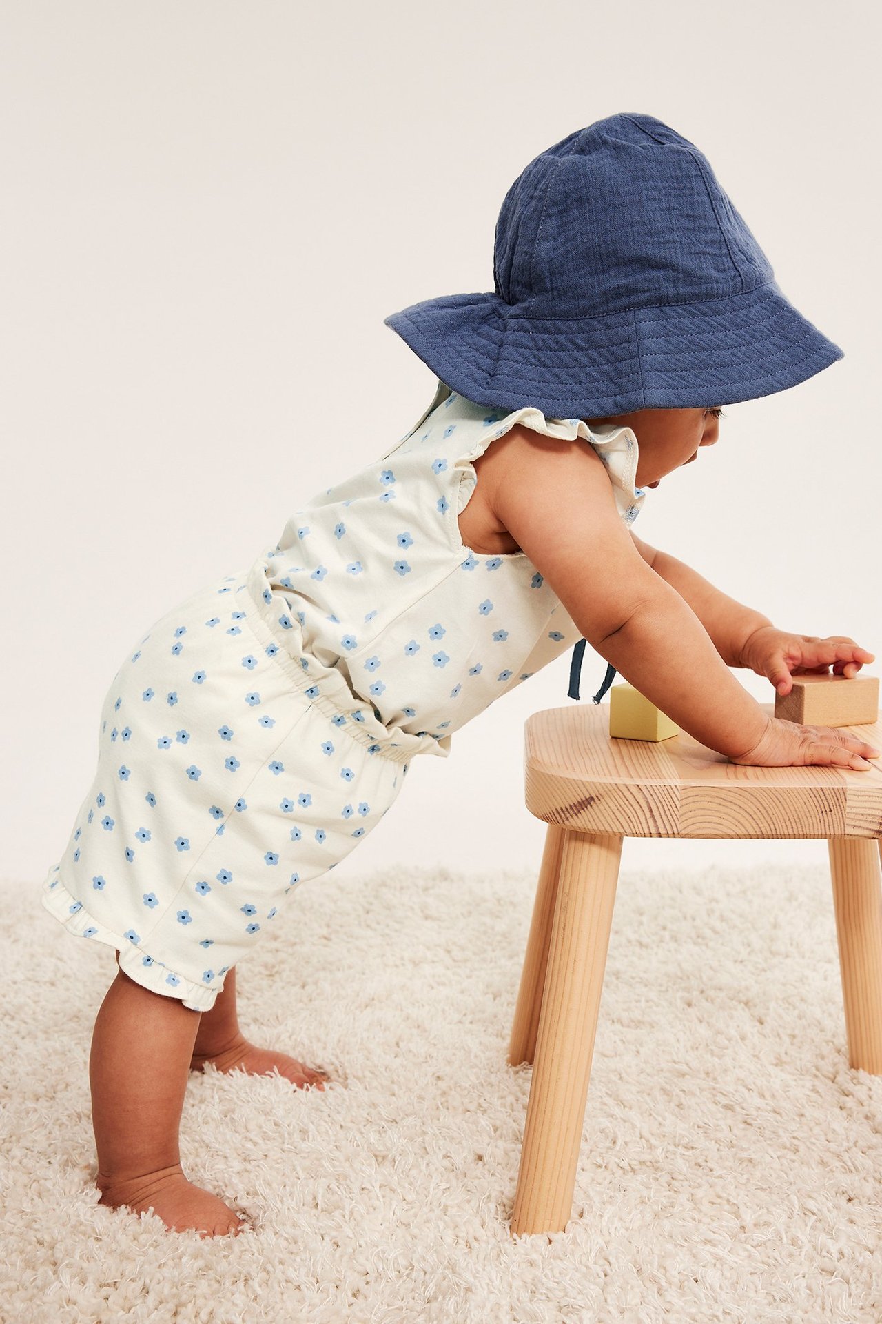 Widok z boku dziecka w białych szortach w kwiaty z falbankami i niebieskim kapeluszu, opierającego się o stołek.