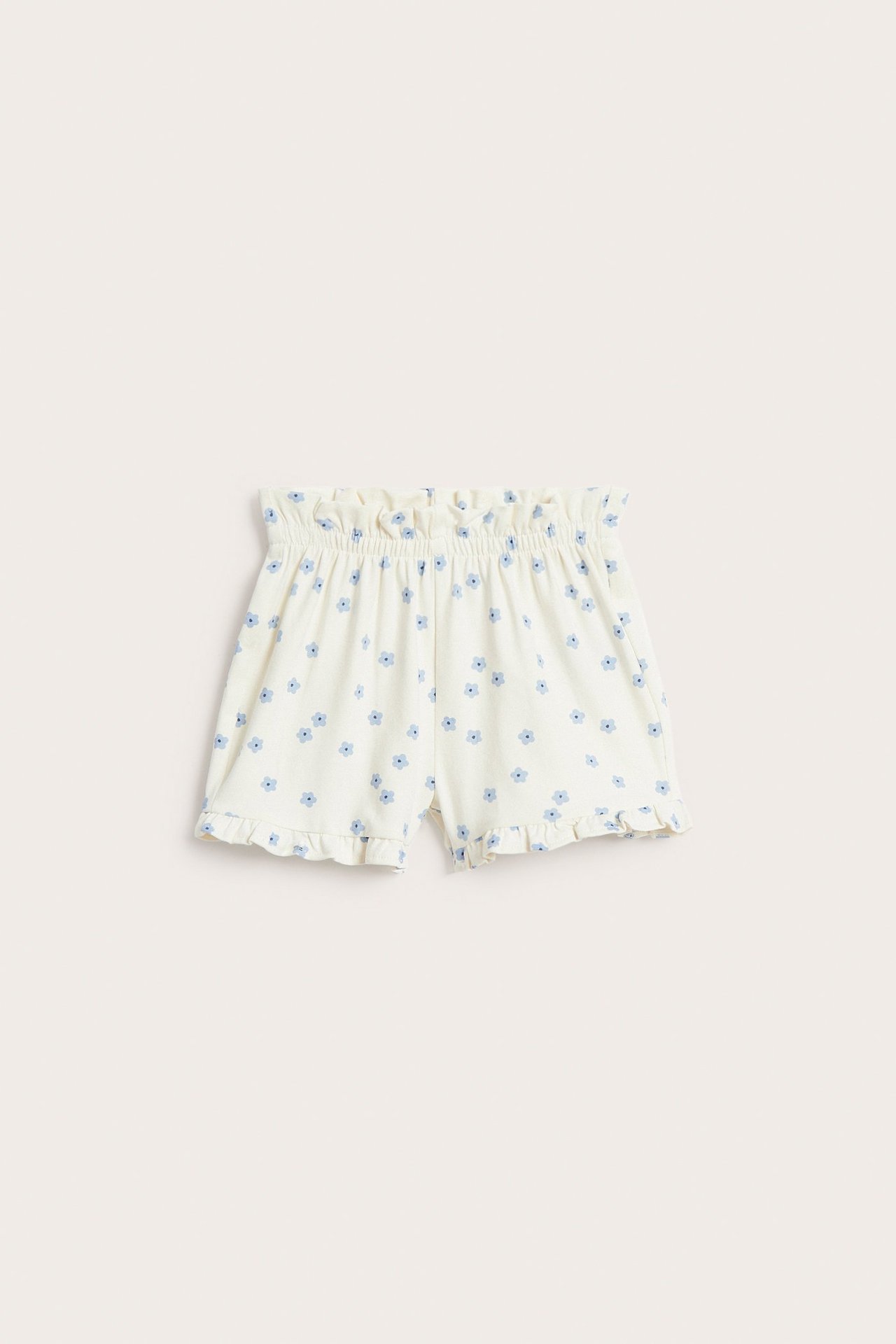 Forfra, baby shorts i offwhite med blått blomstermønster, volang i midjen og volang langs benåpninger.