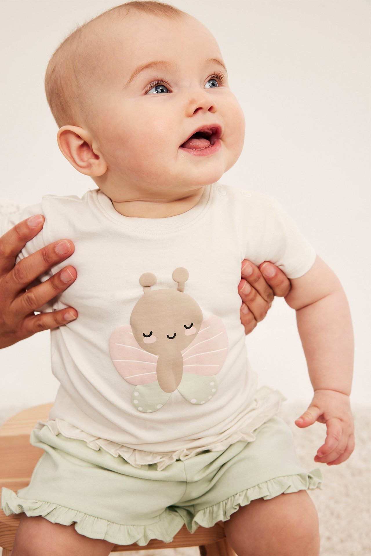 Framsida: Baby bär ljusgröna volangshorts och krämfärgad t-shirt med fjärilsapplikation, hållen av vuxna händer.