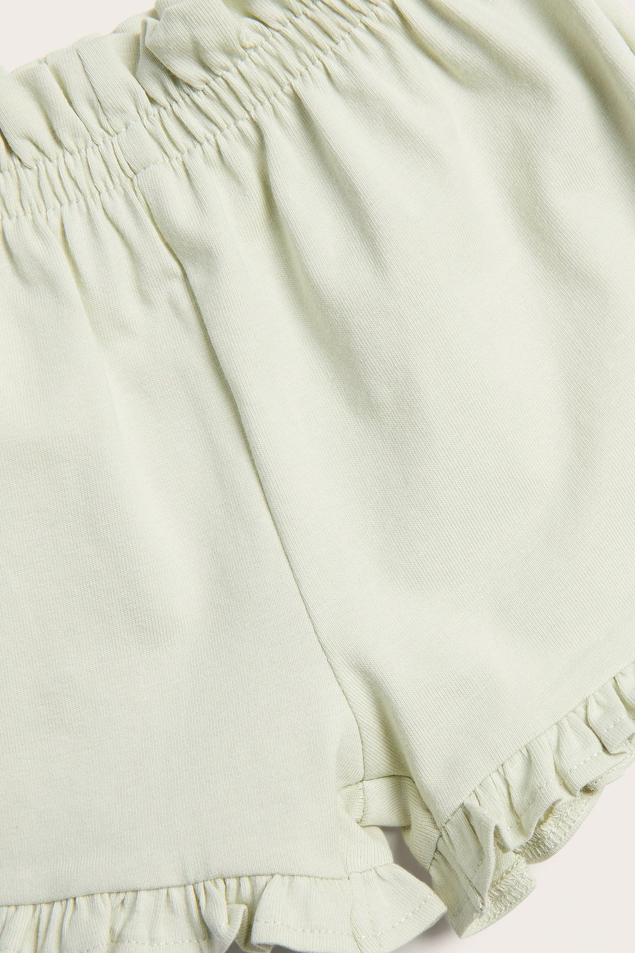 Nærbilde av lys grønne jerseyshorts med rynket strikk i midjen og volanger ved benåpningene.