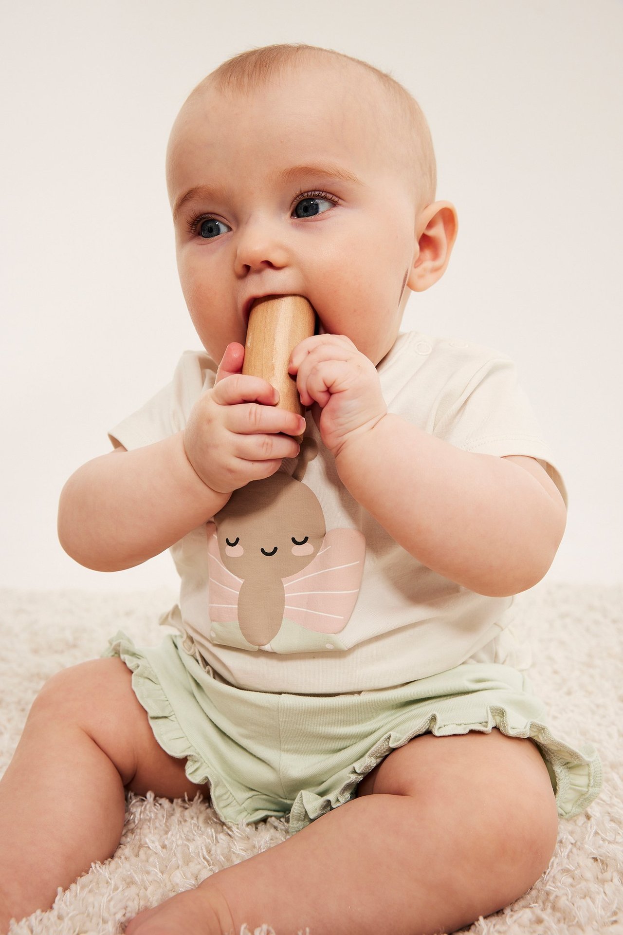 Widok z przodu: niemowlę w jasnozielonych bawełnianych szortach z falbankami i koszulce, trzyma drewnianą zabawkę.