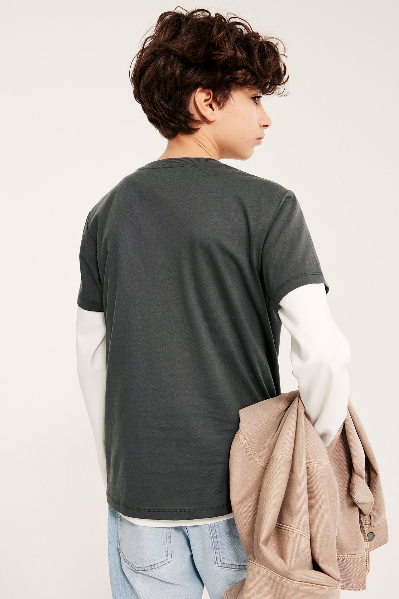 Dziecko w czarnym T-shircie na białej bluzce z długim rękawem, jasnoniebieskich jeansach, trzyma brązową kurtkę.