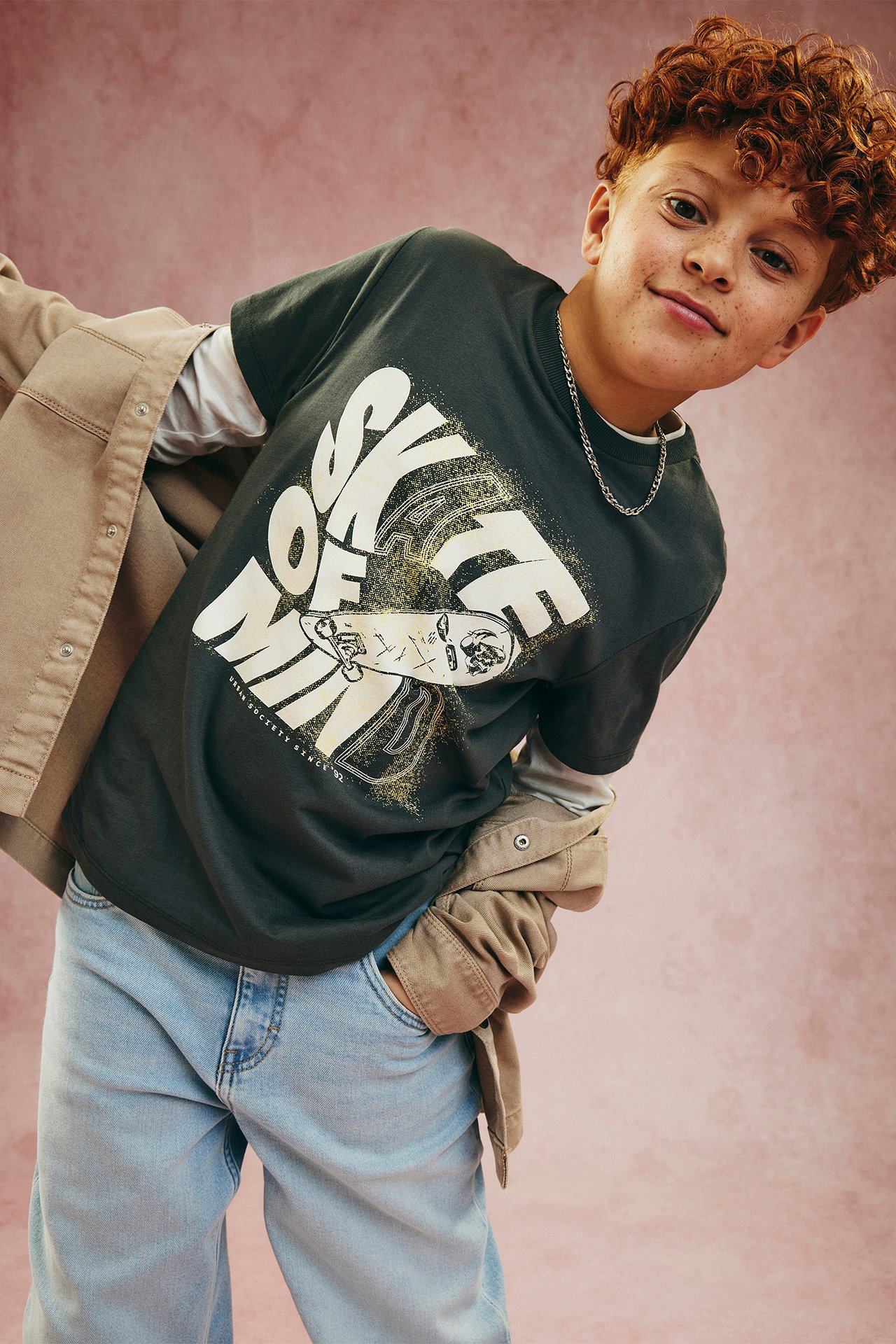 Kille bär svart T-shirt med "SKATE OF MIND"-tryck, ljusgrå topp, beige jacka, ljusblå jeans.