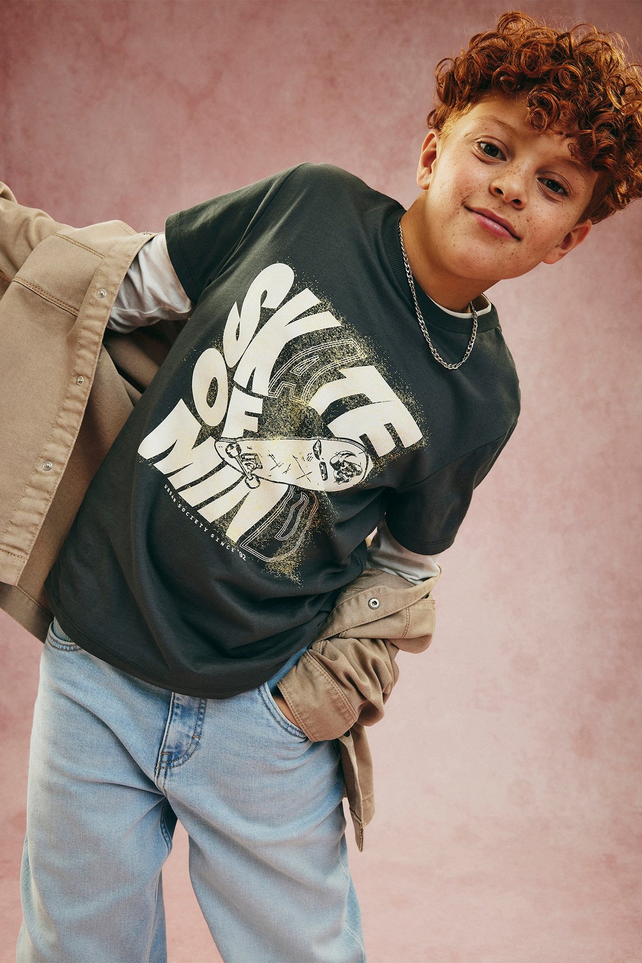 Dziecko w czarnym T-shircie z nadrukiem „SKATE OF MIND”, jasnoszarym topie, beżowej kurtce, jasnoniebieskich jeansach.