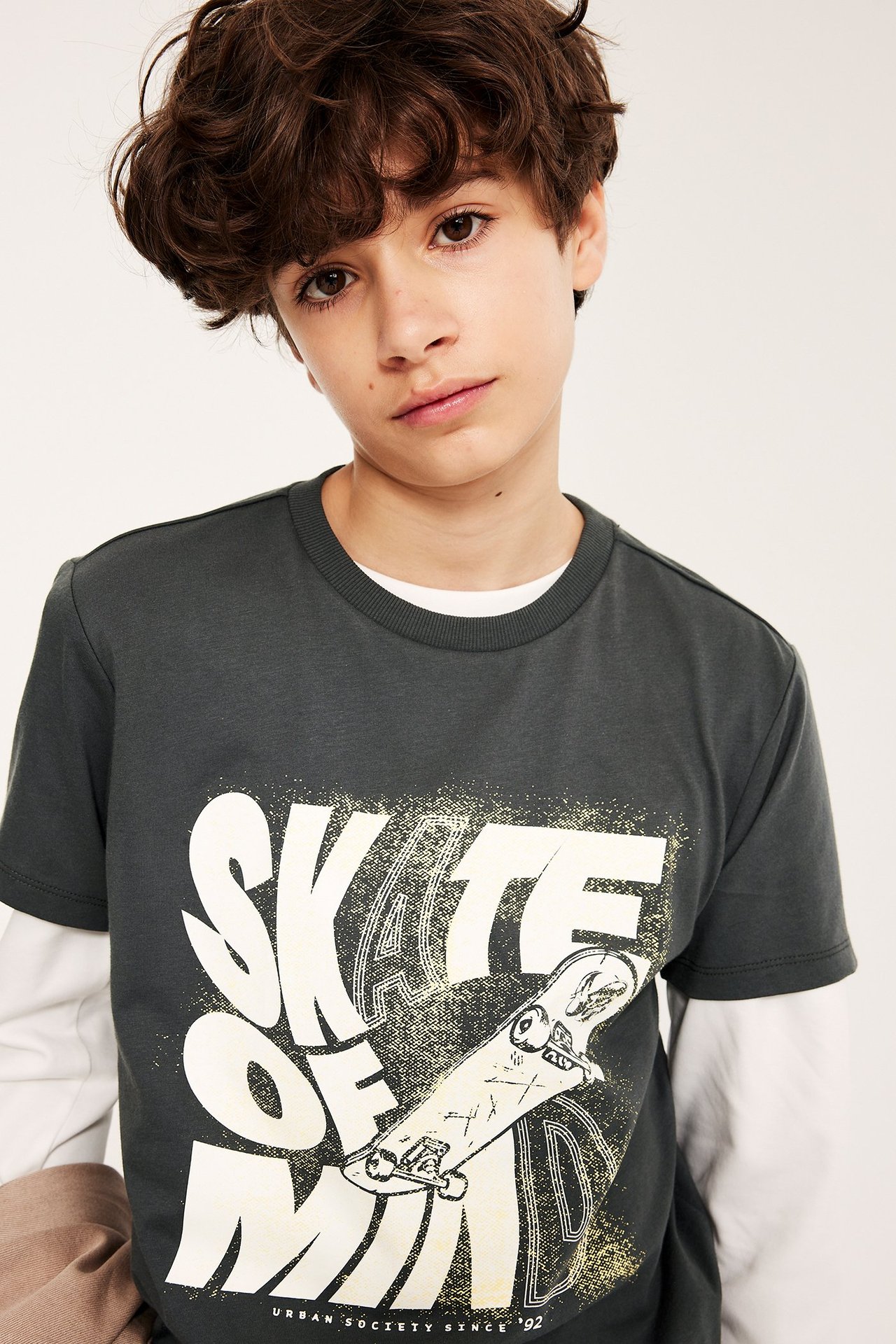 Lapsi pukeutuu mustaan puuvilla T-paitaan 'SKATE OF MIND' -printillä, valkoisen pitkähihaisen paidan päällä.