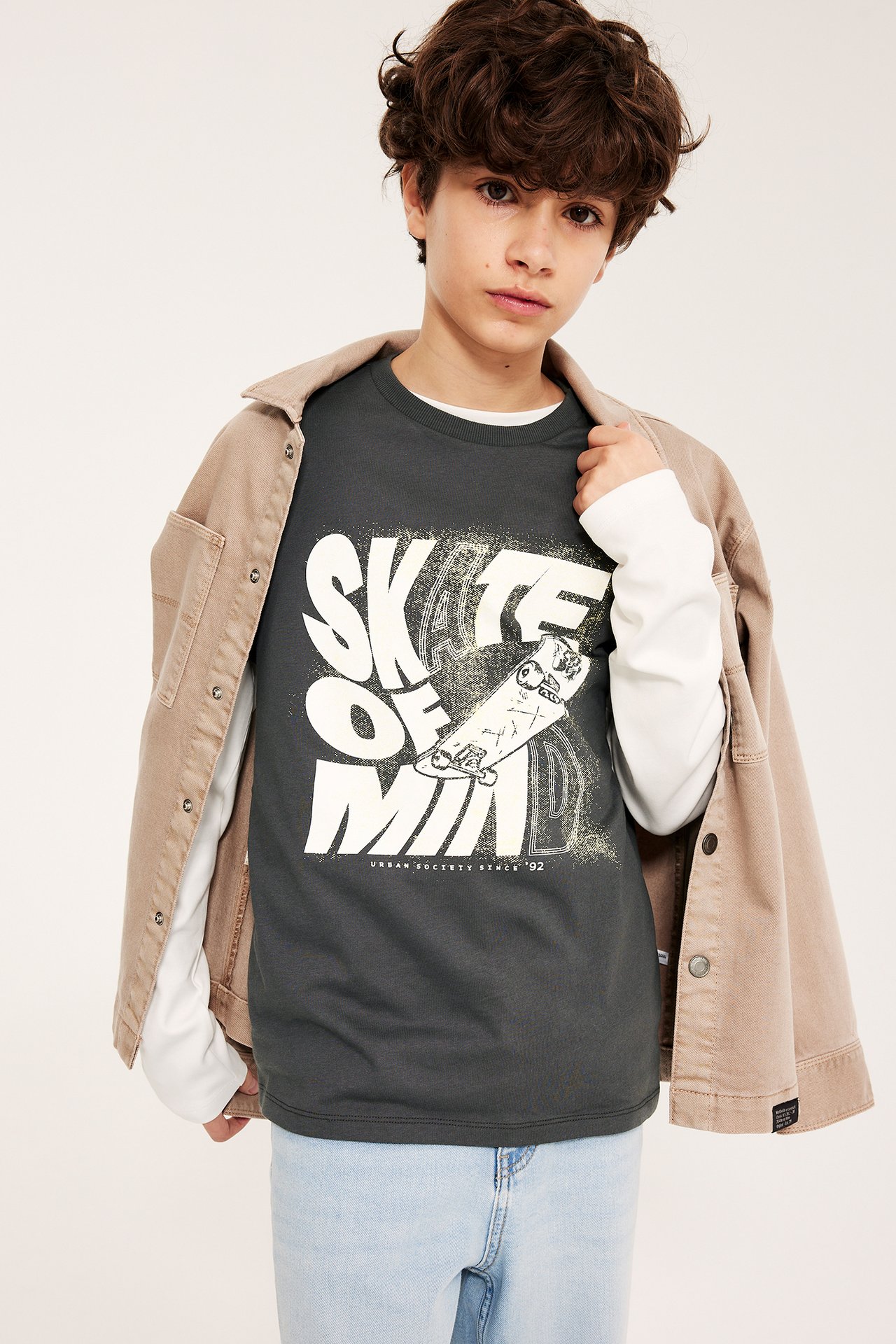 Widok z przodu: Chłopiec w czarnej koszulce z nadrukiem, białym longsleeve, beżowej koszuli, jasnych dżinsach.