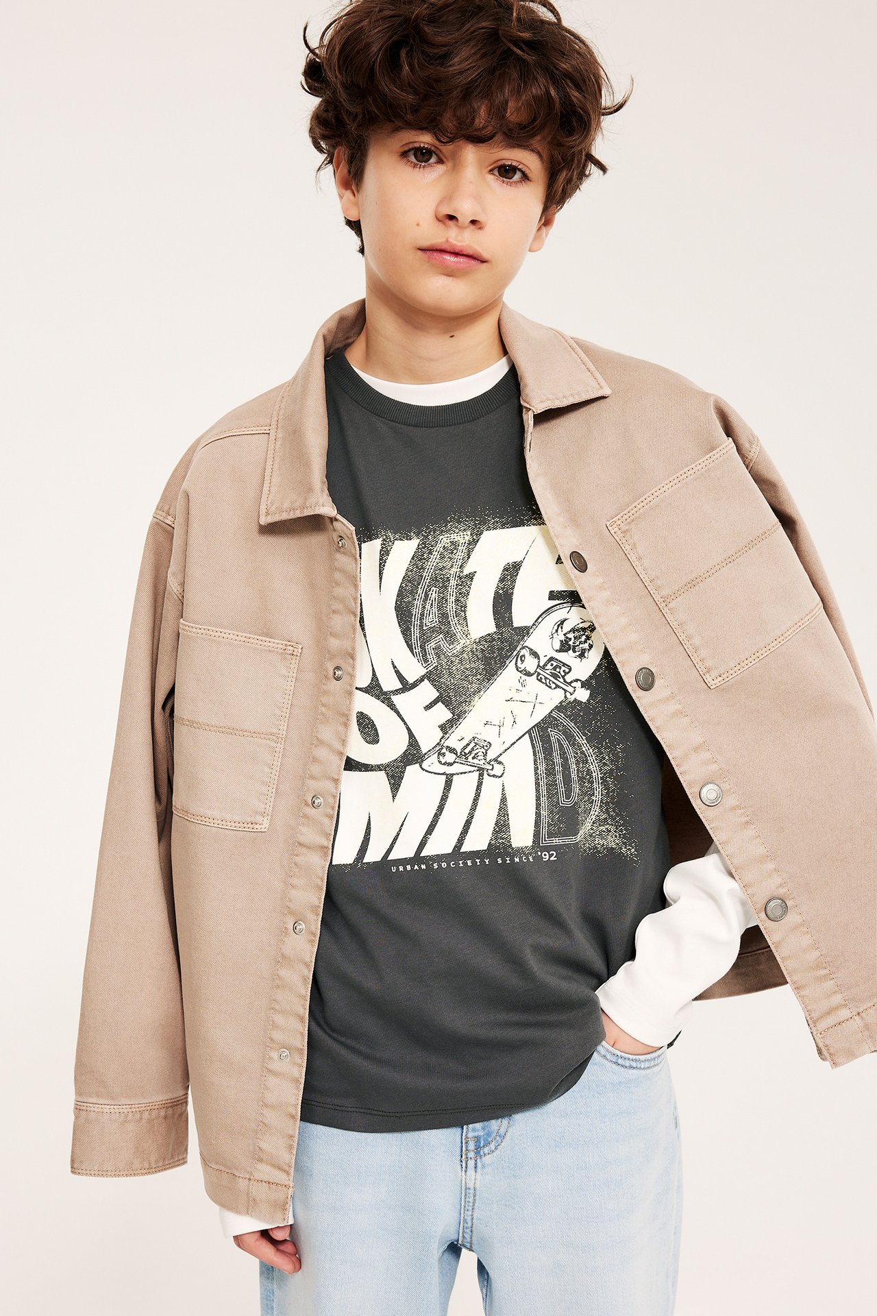 Frontutsikt: gutt i svart T-skjorte med hvitt skateboardtrykk, beige jakke og lyseblå jeans.