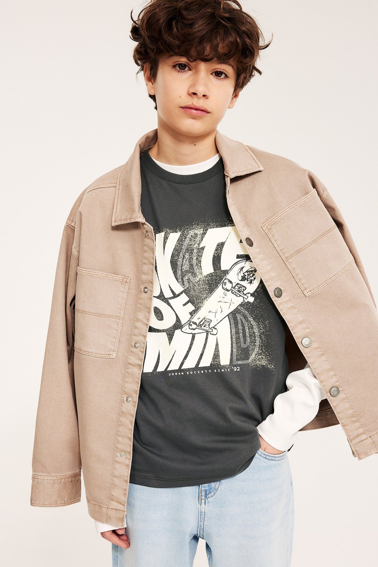 Edestäpäin: poika mustassa t-paidassa valkoisella skeittipainatuksella, beigen takin ja vaaleansinisten farkkujen kanssa.