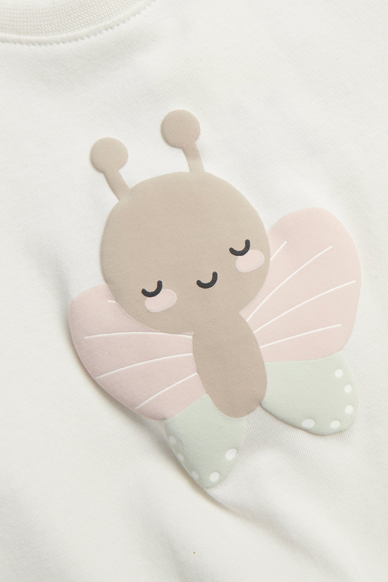 Lähikuva: luonnonvalkoinen vauvan t-paita, jossa beige, vaaleanpunainen ja vihreä kohoperhosprintti.