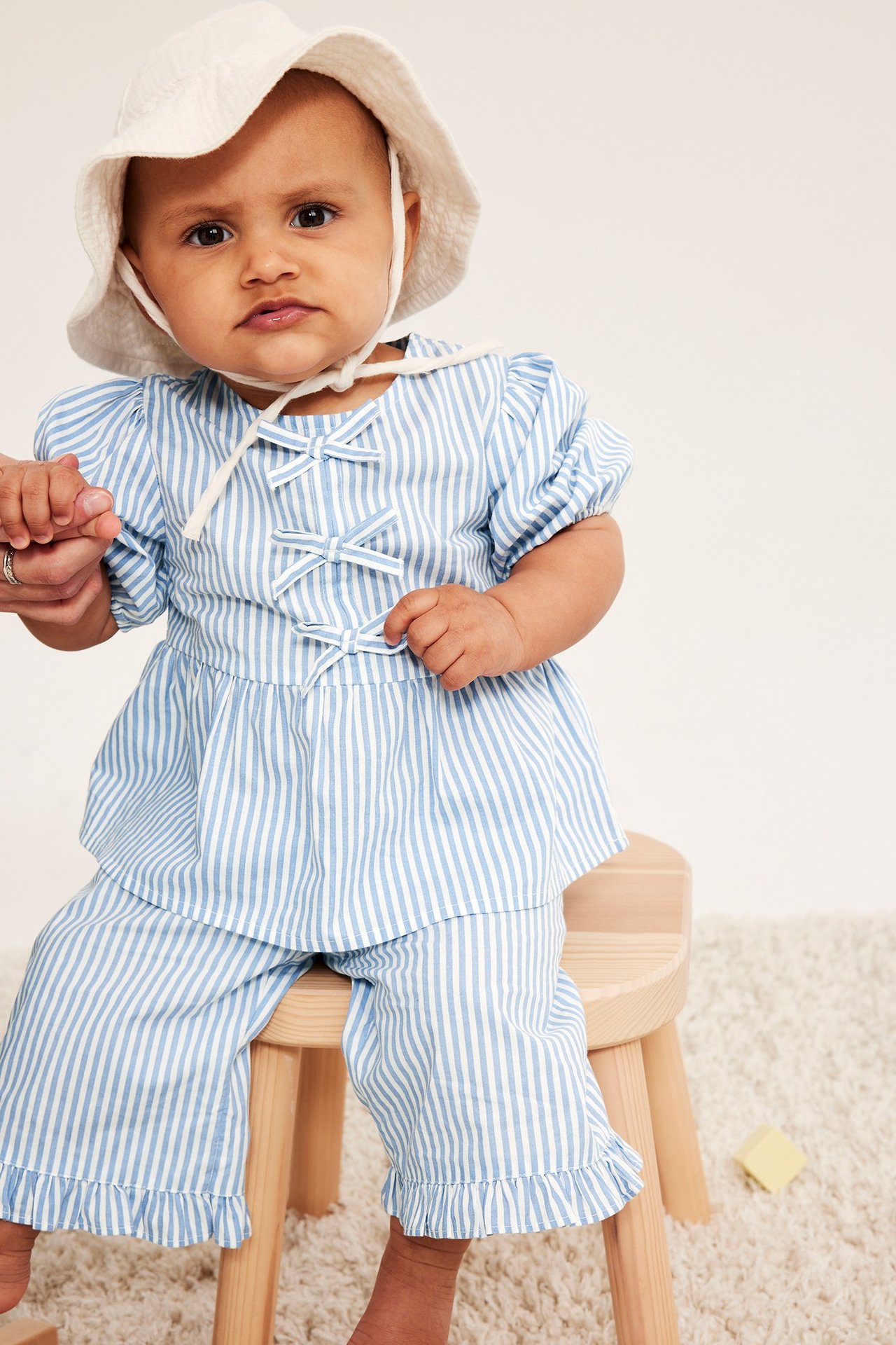 Framsida: Baby i ljusblårandiga poplinbyxor med volanger, matchande topp med rosetter och vit hatt.