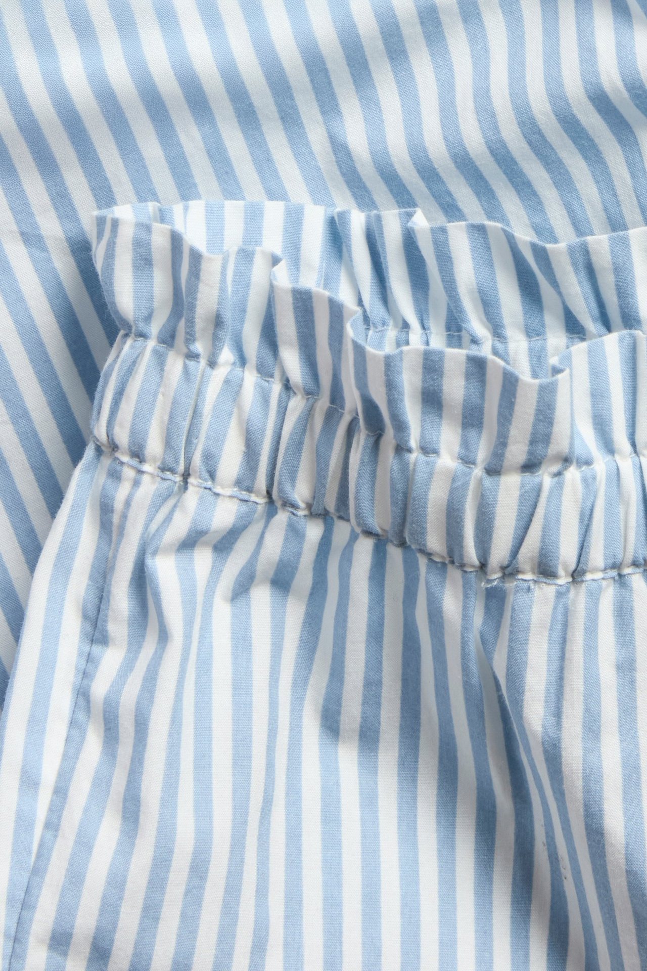 Nærbilde av lysblå og hvitstripete babybukser i poplin med elastisk midje og volanger.