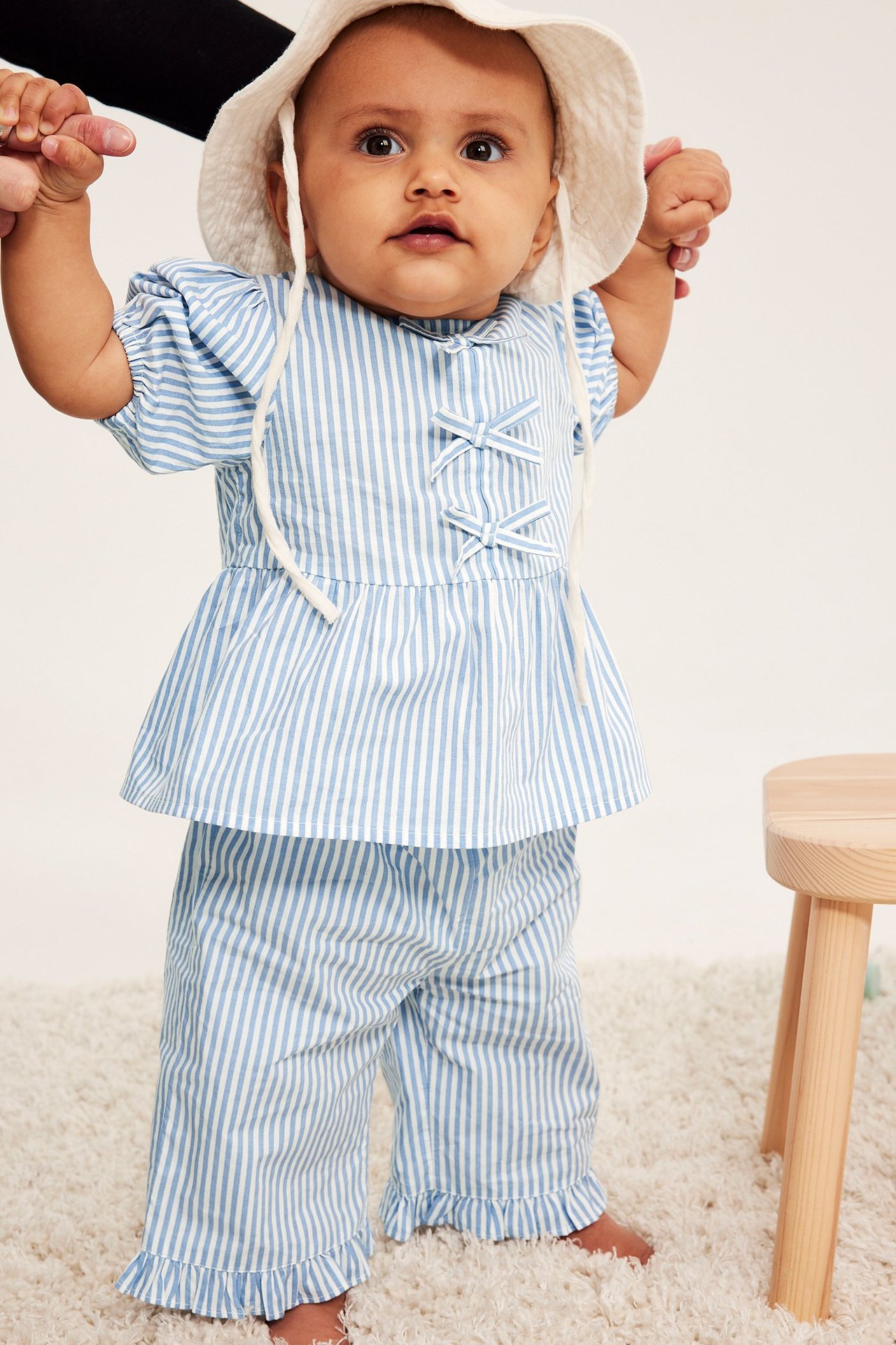 Framifrån: baby i ljusblårandiga volangpoplinbyxor, matchande topp och vit hatt.