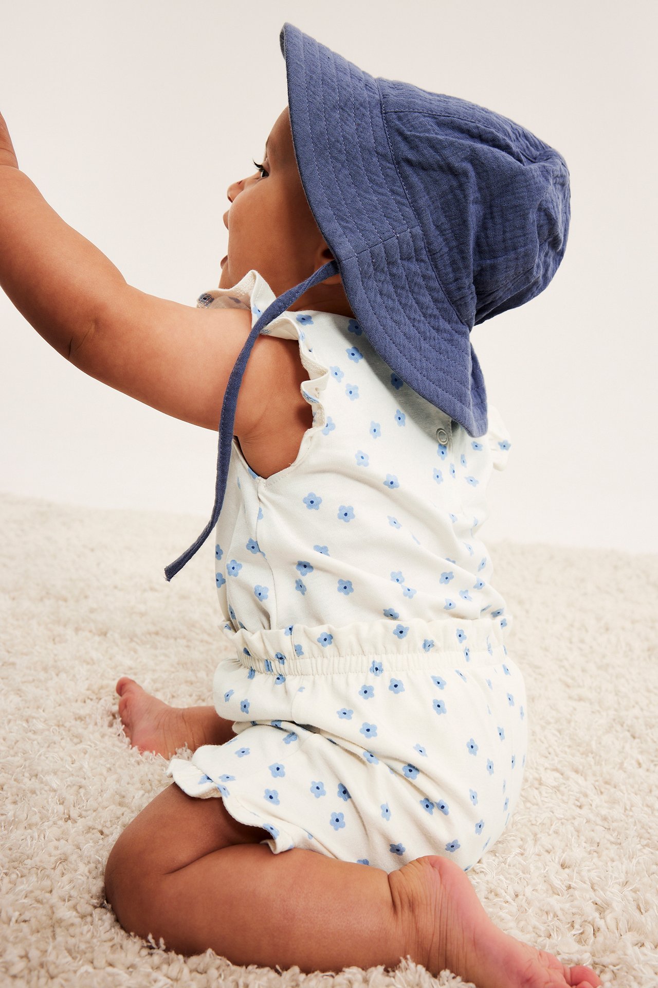 Sidebilde av baby i offwhite blomstret ermeløs body med volanger og blå vattert bøttehatt.